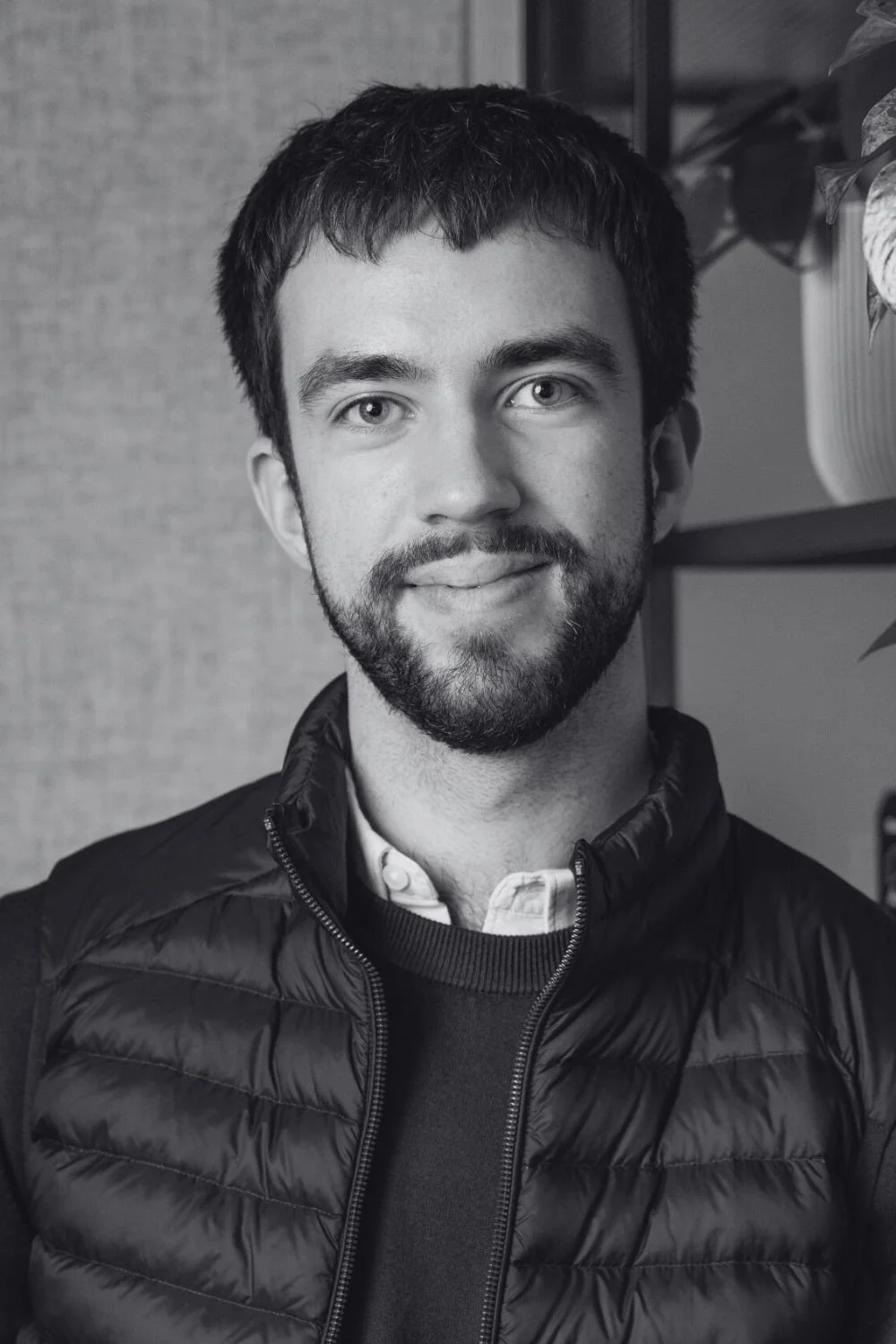 Black and white photo of Ryan, a young man with short hair, a beard, and wearing a Patagonia jacket with the Cambridge Future Tech logo, standing outdoors with blurred trees and buildings in the background.