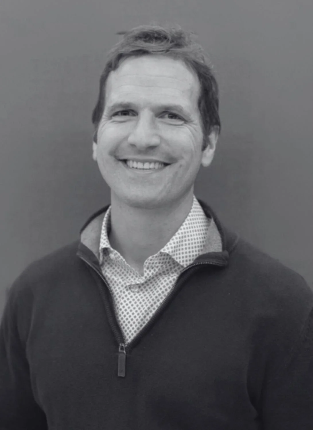A black and white photo of Tim, a smiling man with short, tousled hair, wearing a patterned button-up shirt and a zippered dark jacket, standing in front of a plain background.