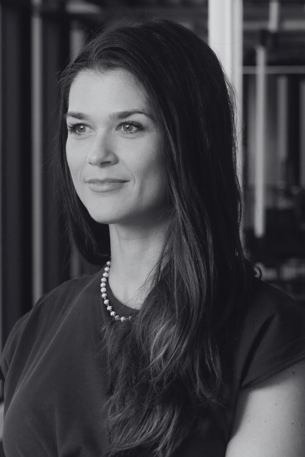 A black and white photo of a Katie with dark hair and light eyes, smiling slightly, wearing a zip-up jacket and a turtleneck sweater, standing in front of a bookshelf.
