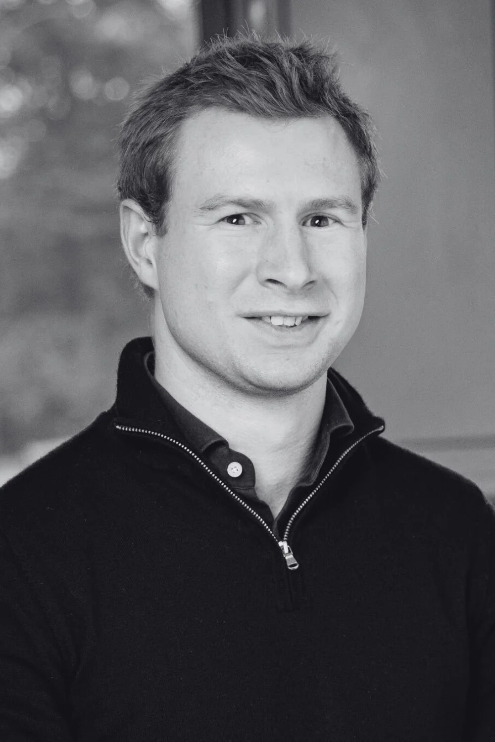 Black and white portrait of Maxim, a young man with short hair, smiling slightly, wearing a Patagonia fleece jacket with logos, leaning against a brick wall.