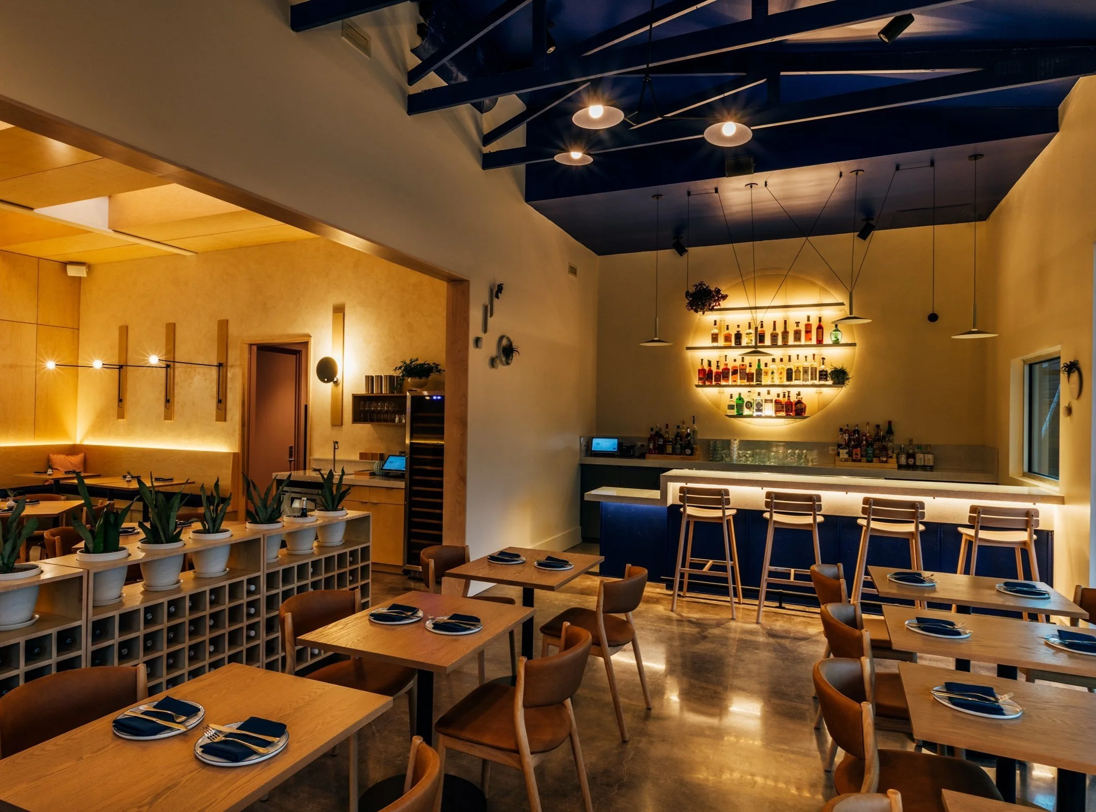 Interior of a modern restaurant or bar with wooden tables set with black napkins and cutlery, a bar area with chairs, and warm ambient lighting.