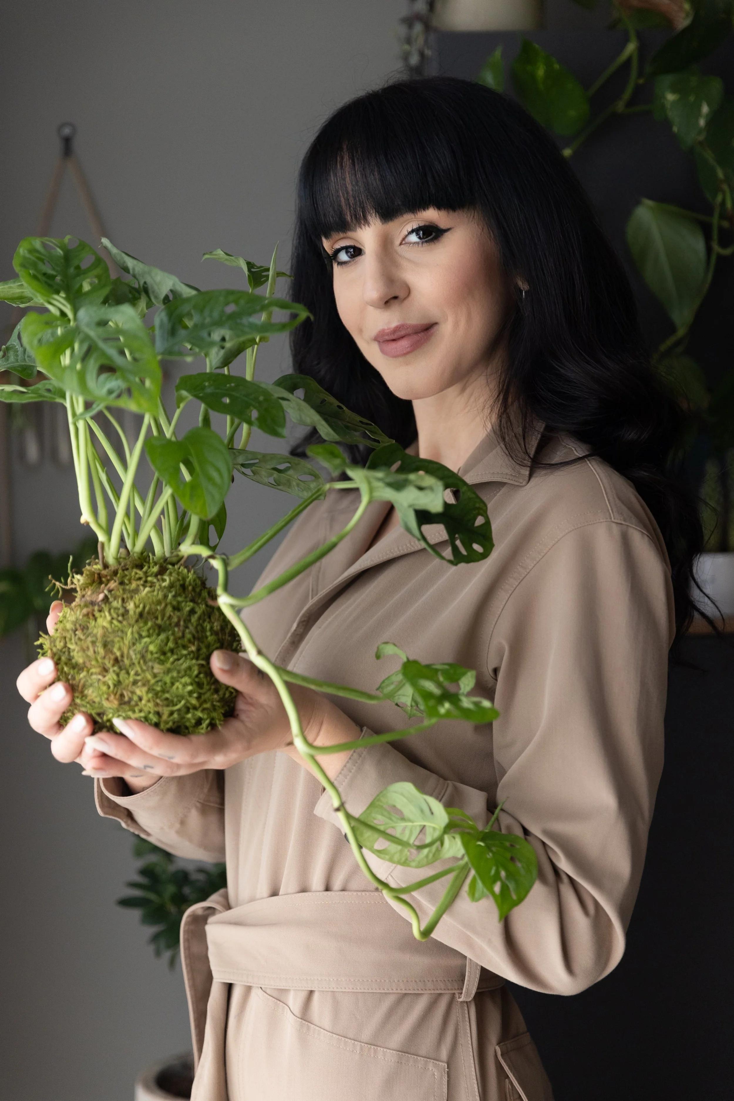 Una donna con capelli neri e boccoli tiene in mano una pianta di Monstera in un ambiente interno con piante sullo sfondo.