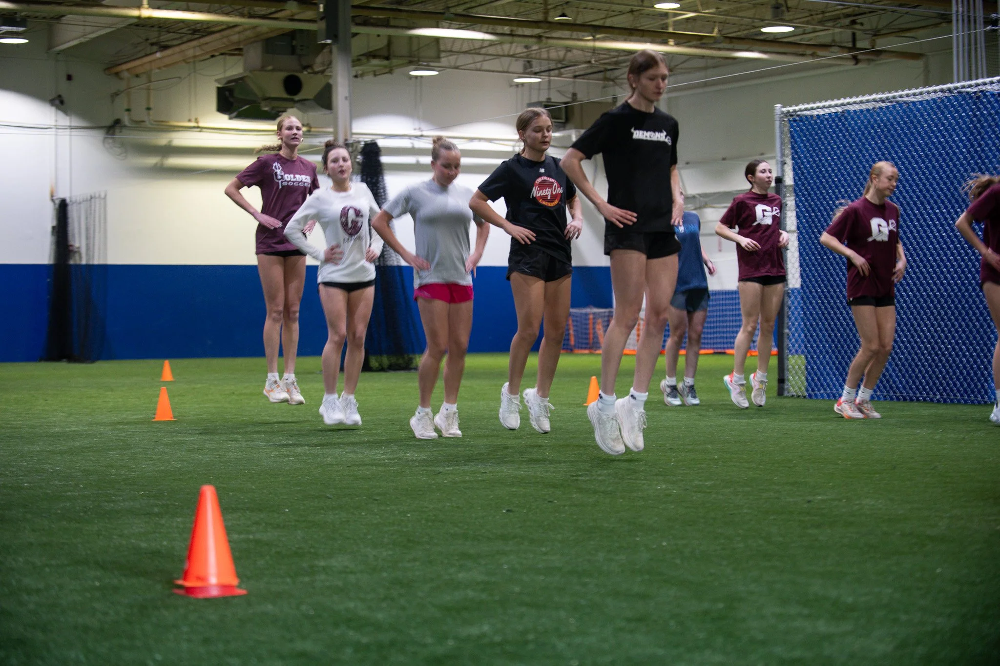 A group of young women participating in a fitness or sports training session indoors on a turf field. They are running between orange cones in athletic attire at Golden Sports Performance in the Applewood neighborhood of Lakewood, Colorado.