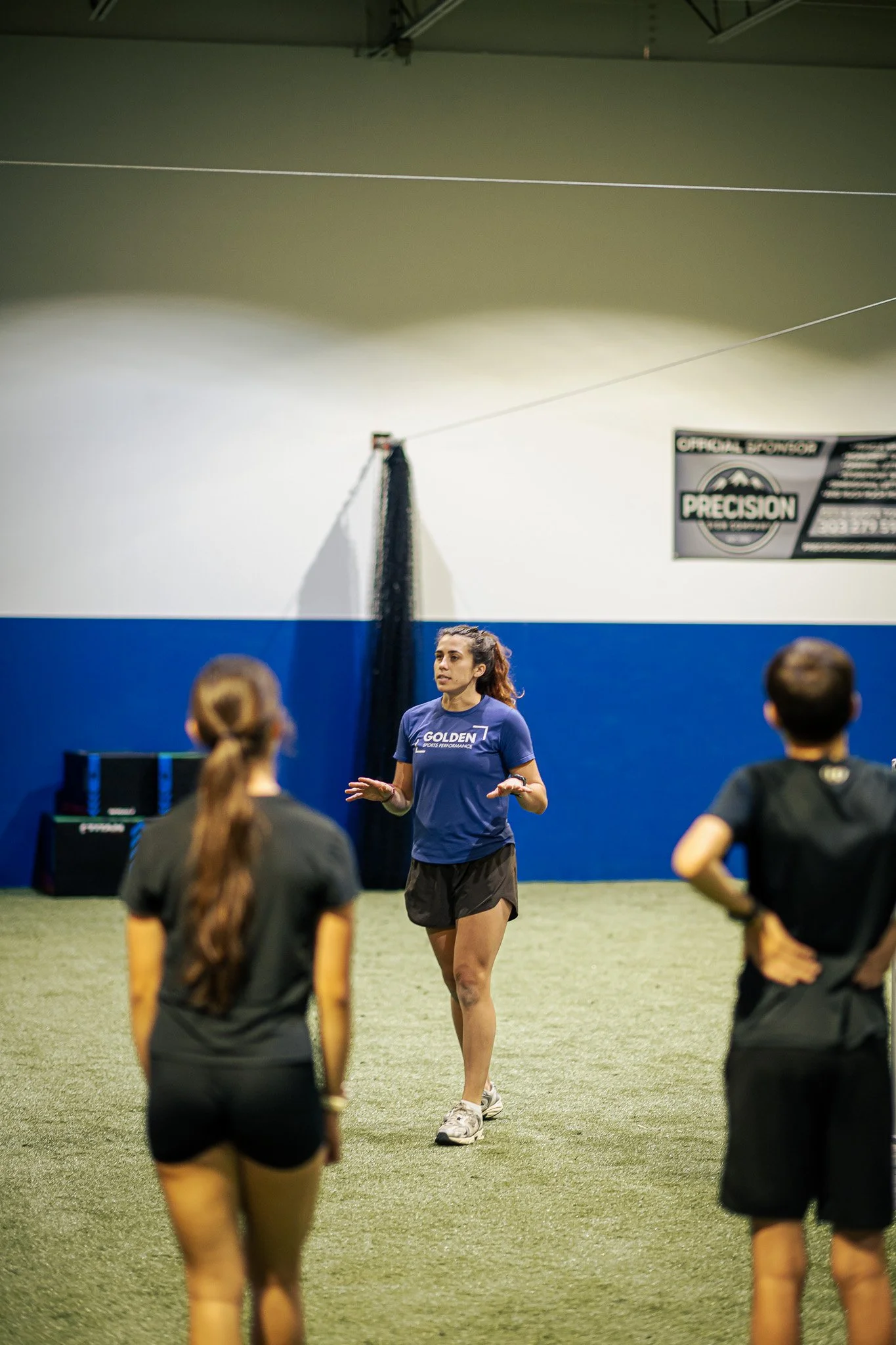 Coach Kaylee Phillips giving instructions to a group of young athletes inside an indoor training facility. The background shows a sign that says 'Precision' and some stacked equipment at Golden Sports Performance in Lakewood, Colorado