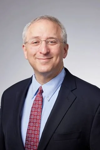 A middle-aged man with glasses, graying hair, wearing a dark suit, light blue shirt, and patterned red tie, smiling at the camera.