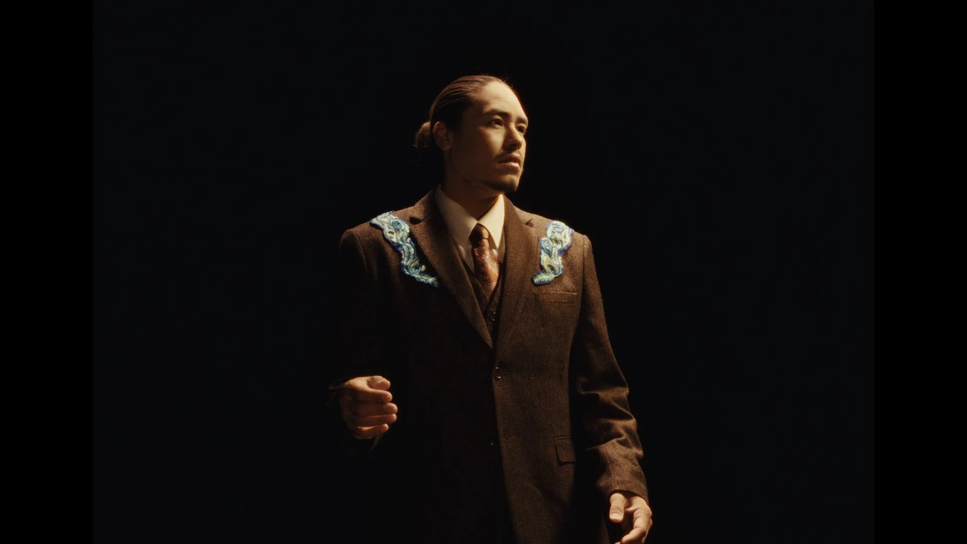 A man in a brown suit with embroidered blue and white details on the shoulders, standing against a dark background, looking thoughtfully into the distance.