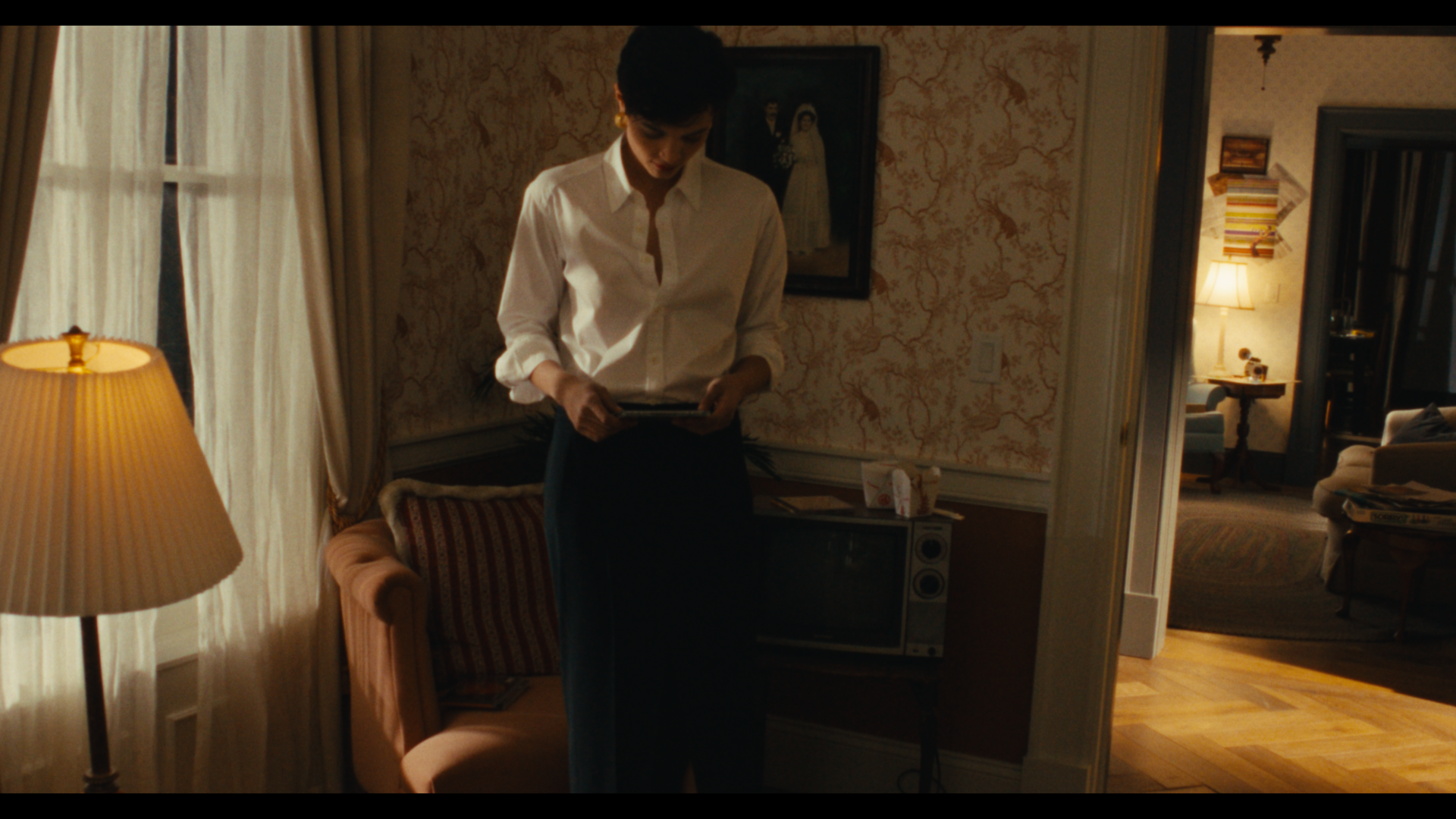 A woman in a white shirt and dark pants standing in a warmly lit room, holding a book or folder, with a patterned wallpaper, a framed photo or painting, and a corner with a lamp and furniture visible.