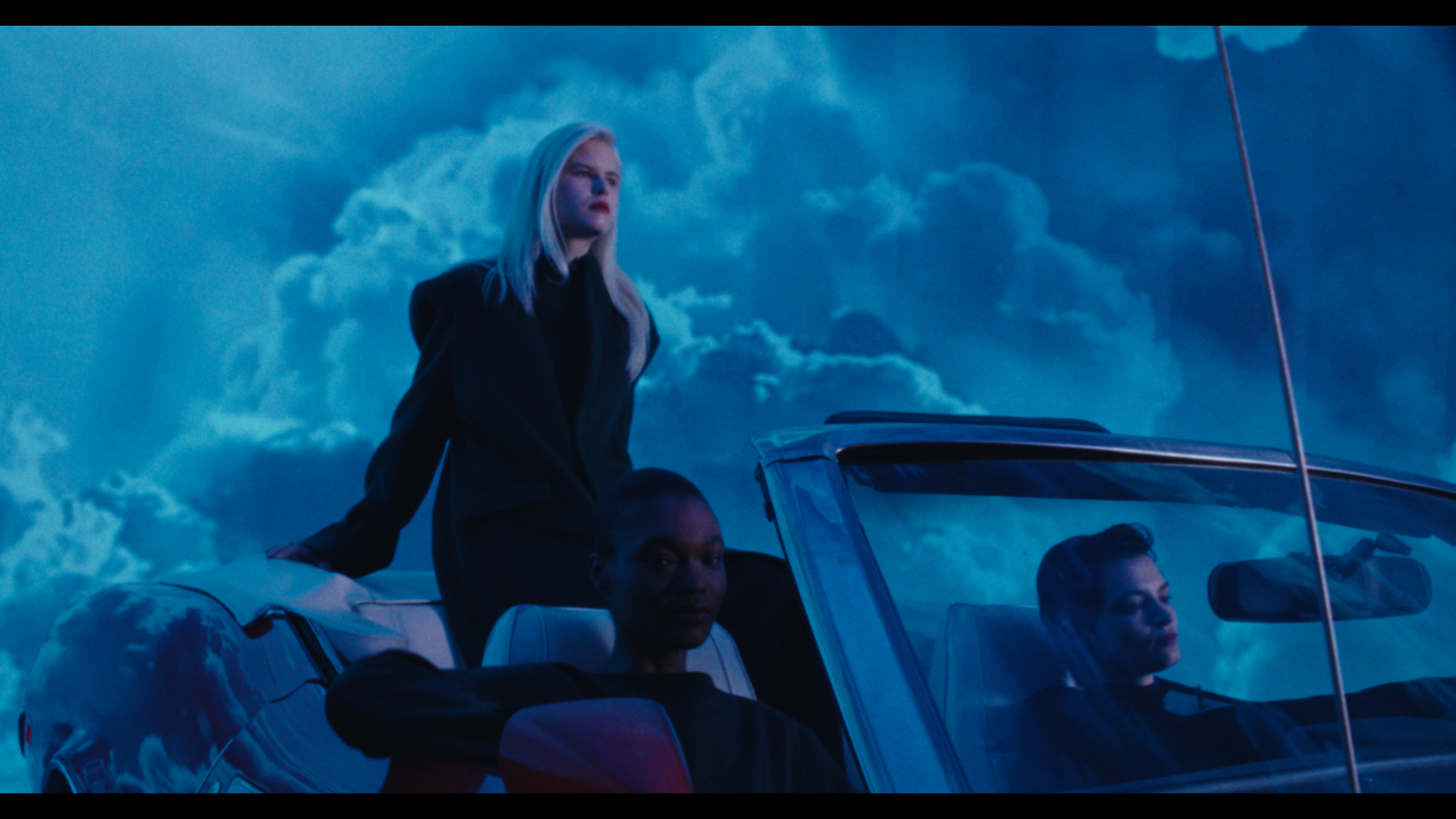 Three women in black clothing riding in a convertible car against a background of dramatic clouds in the sky.
