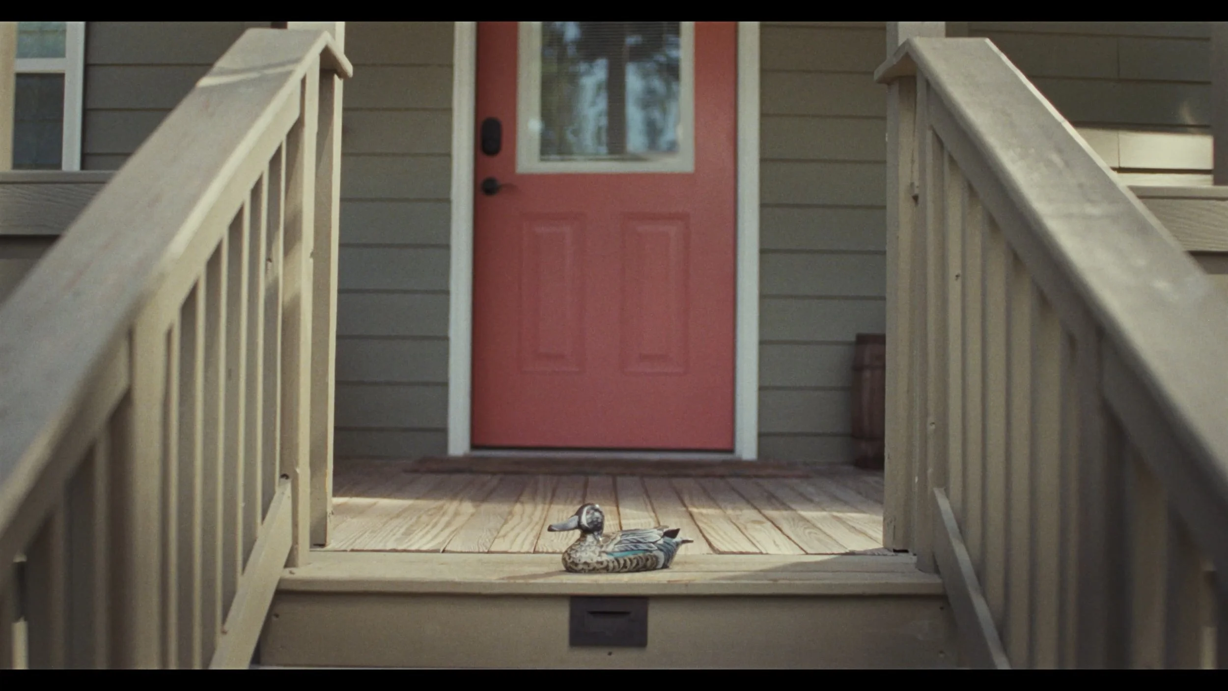A decorative duck figurine on a wooden porch floor in front of a red front door with a small window, beige siding, and two beige railings on either side.