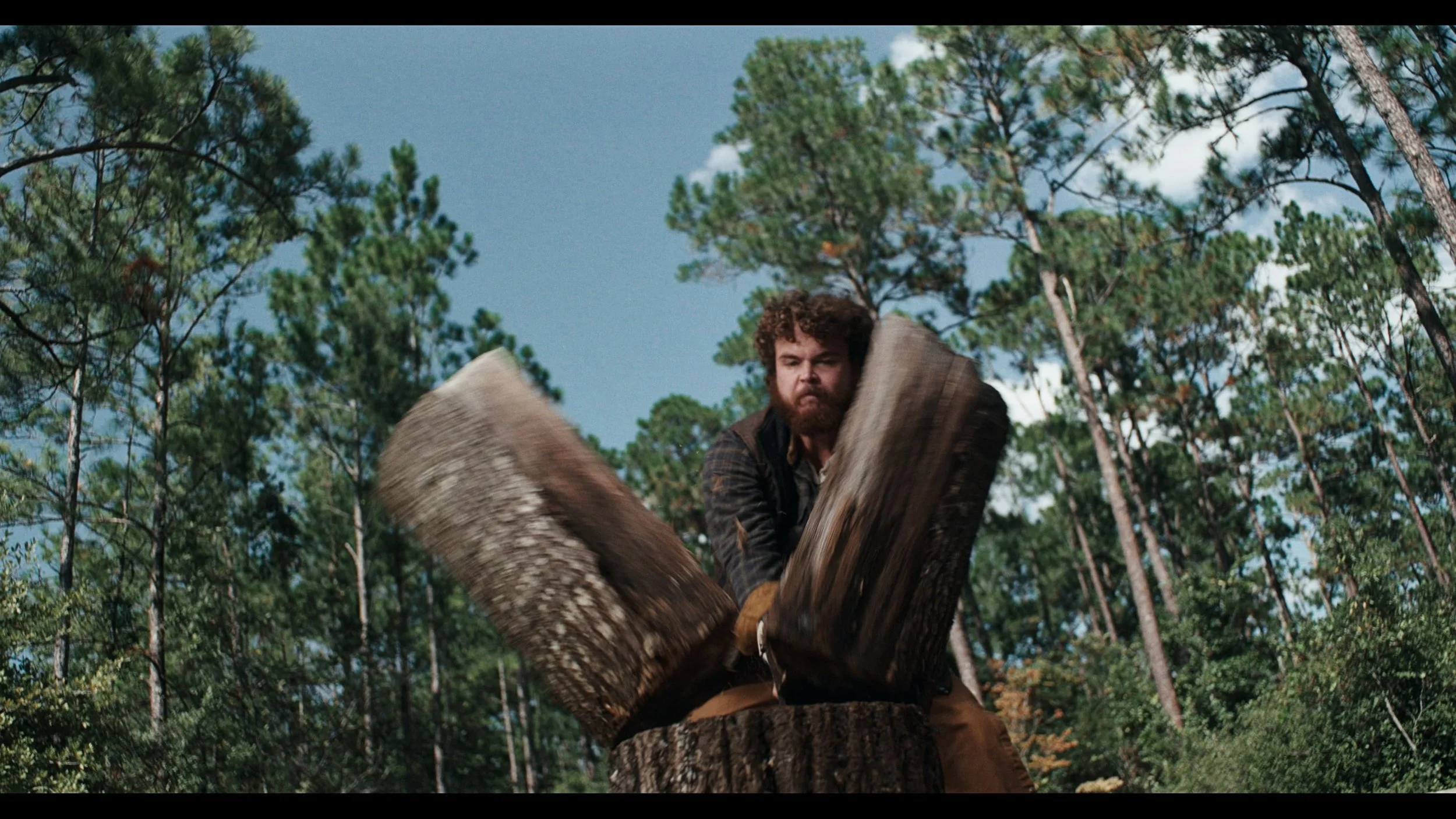 A man with curly hair and a beard splitting a large log with an axe in a forest clearing.