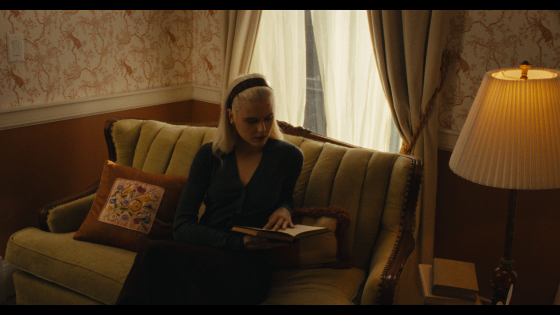 A woman with blonde hair wearing a black headband, sitting on a vintage green sofa in a warmly lit room, reading a book. The room has floral wallpaper, cream curtains, and a side table with a lamp and books.