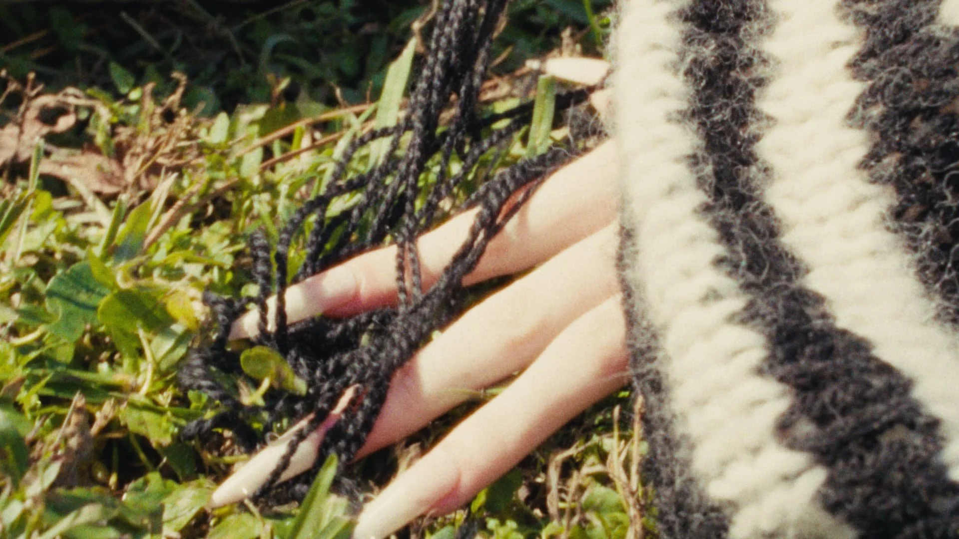 Close-up of a hand with long, pointed, white nails resting on grass and surrounded by black, shiny braids and a black and white knitted fabric.