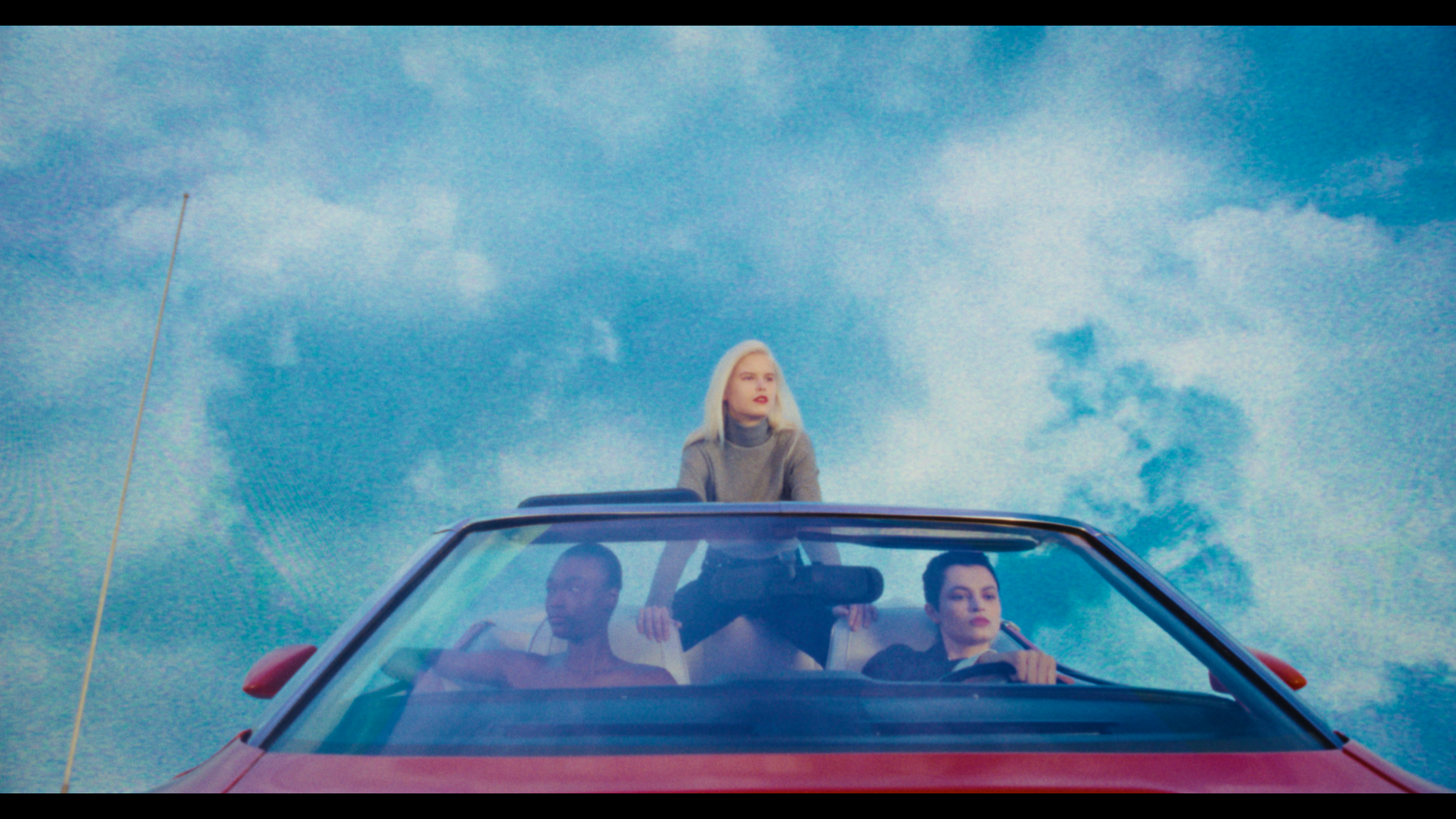 Three young women in a red convertible car driving under a cloudy sky.