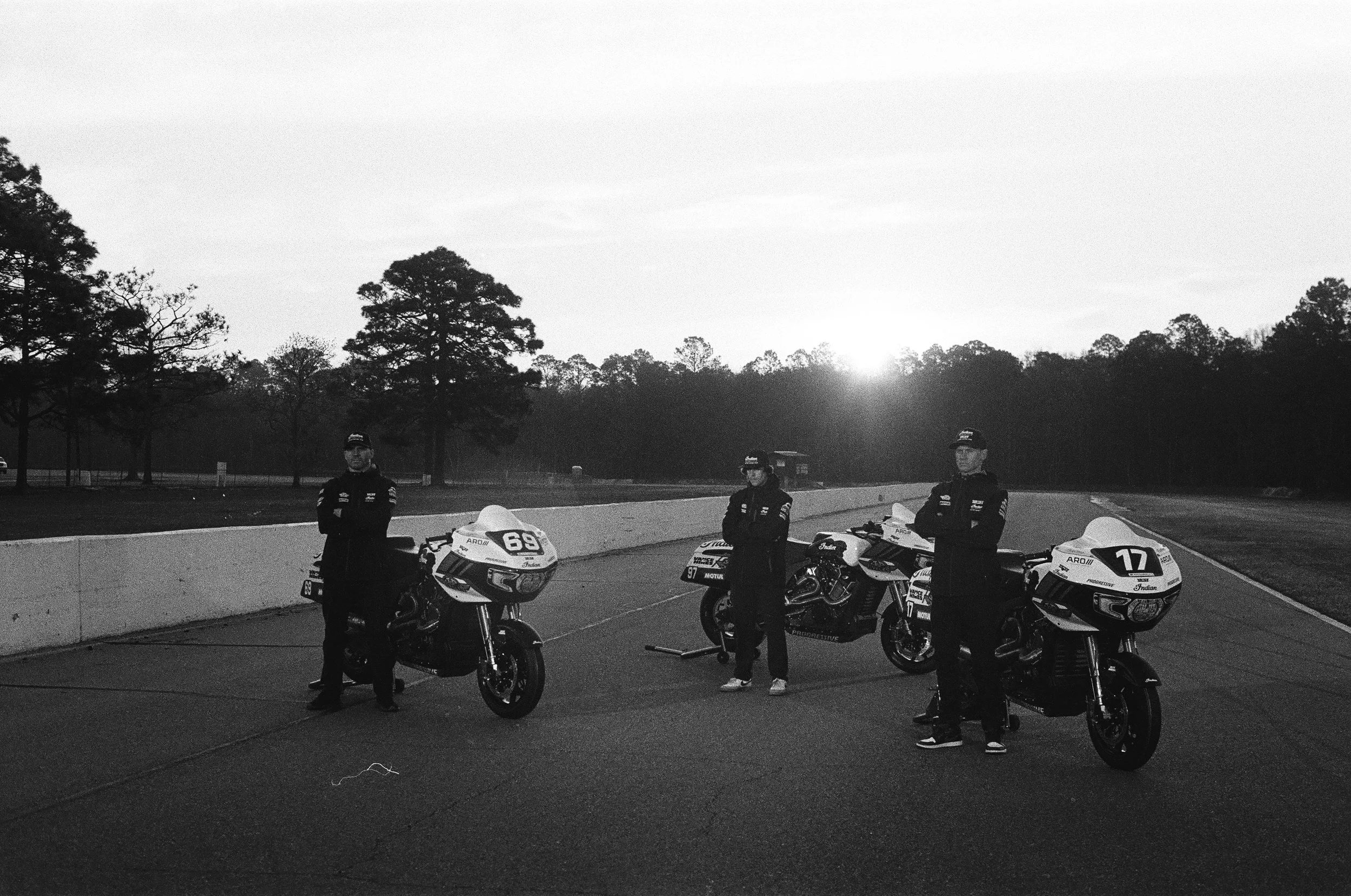 Three race car drivers standing next to their motorcycles on a racetrack at sunset, wearing racing suits and helmets on the Indian Motorcycle racing team, behind the scenes by Mathias White