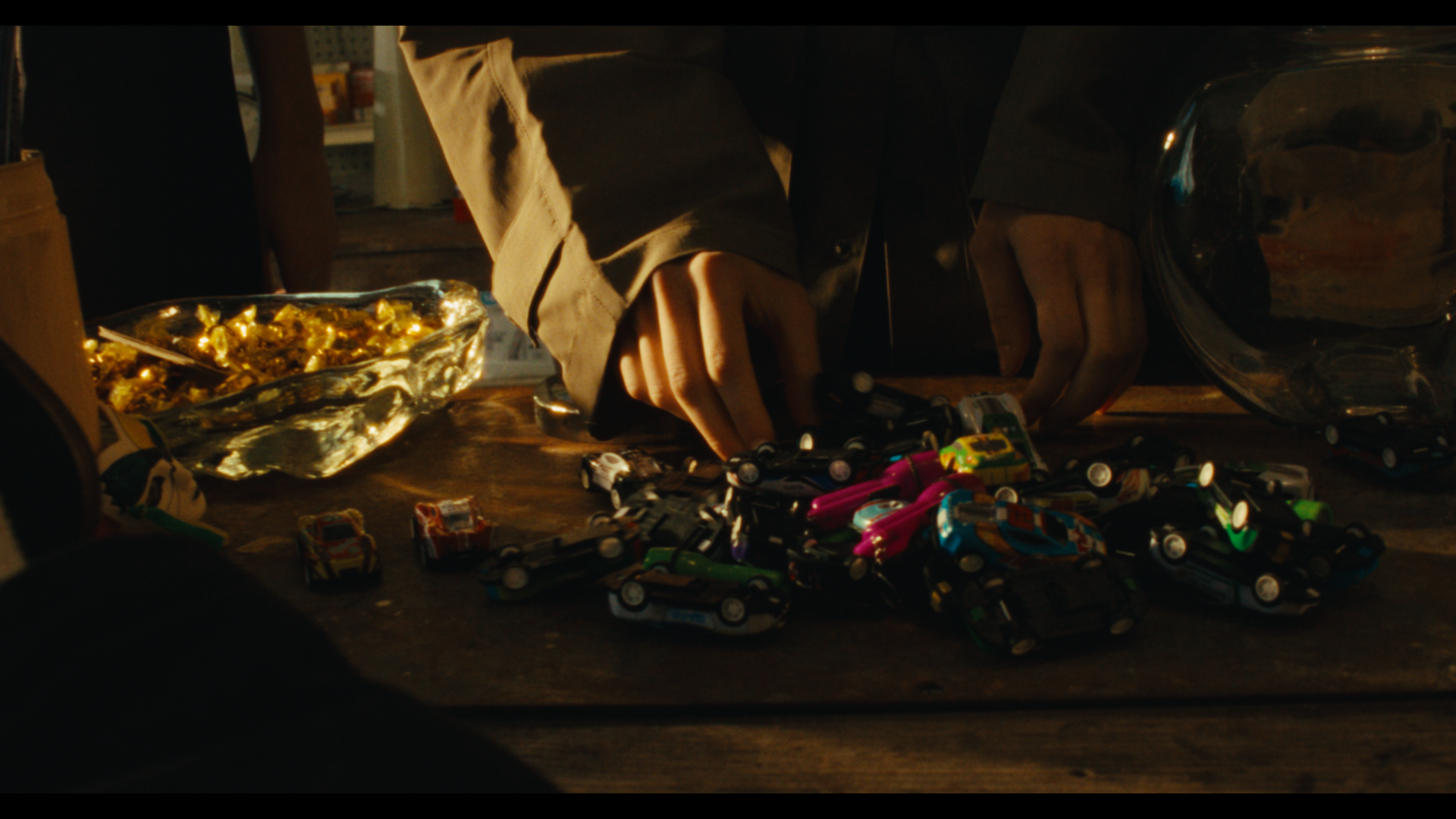 Close-up of hands arranging small toy cars on a wooden surface, with a pile of more toys and a glass bowl nearby in warm lighting.