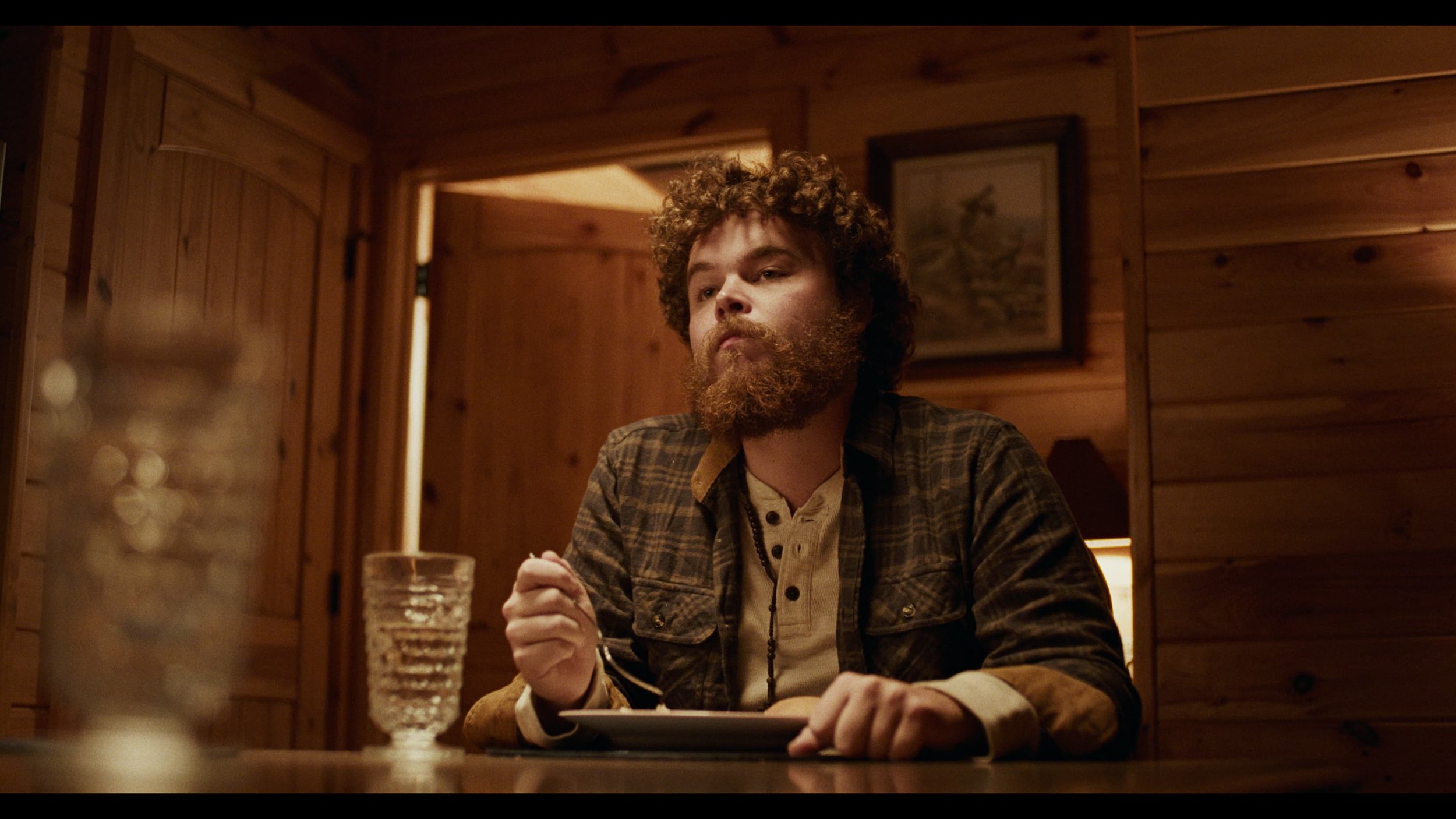 A man with curly hair and a beard sitting at a wooden table in a cozy, wood-paneled room, holding a fork in one hand and a plate in front of him, with a glass of water and a blurred object in the foreground.