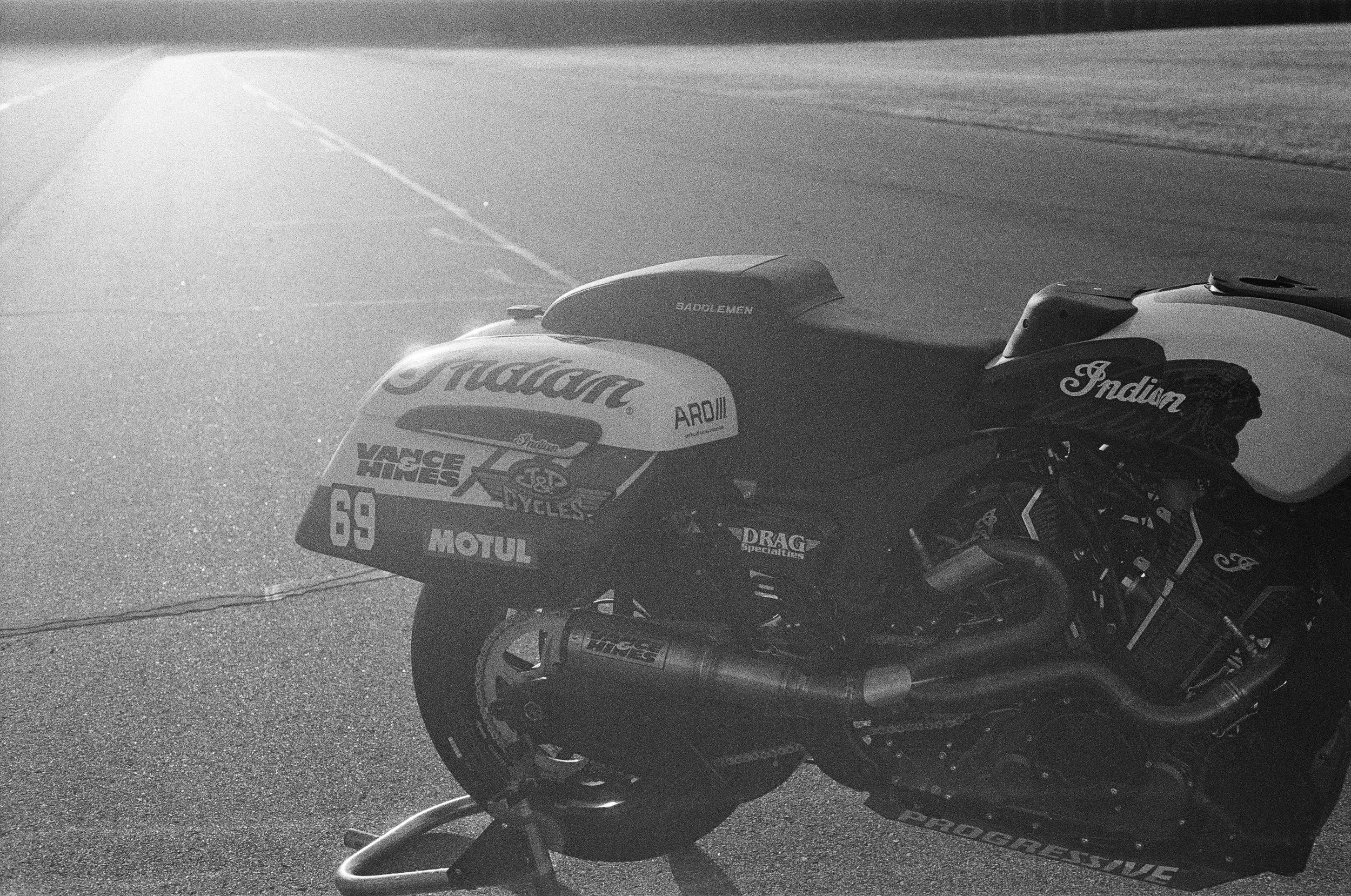 Black and white photograph of a motorcycle with sponsor logos parked on an empty road. Indian Motorcycle Racing Team Behind the scenes by Mathias White