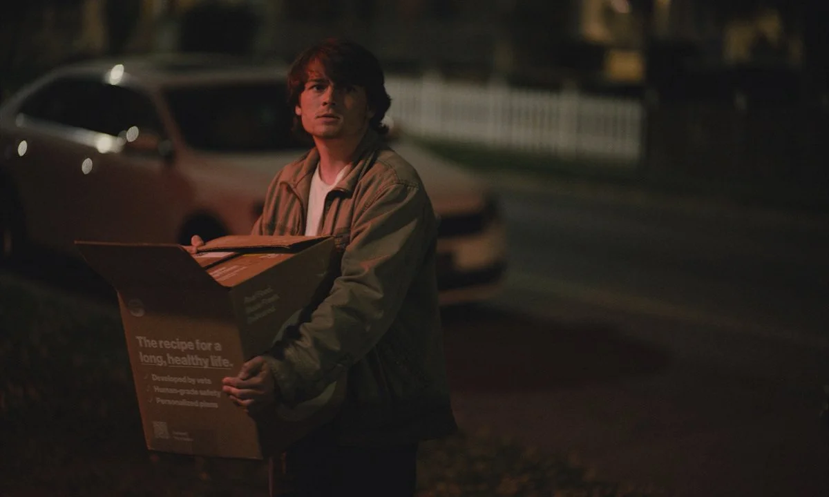 A young man with brown hair wearing a beige jacket is holding a pizza box while standing on a dark street at night. A car is parked behind him.