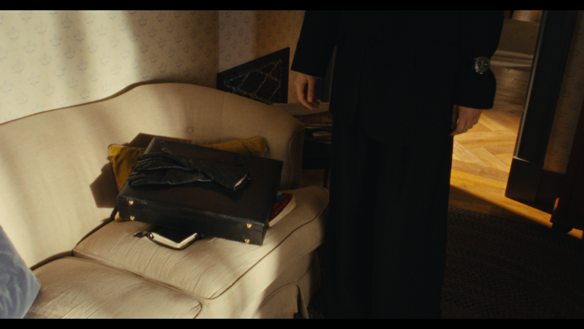 A beige sofa with a black handbag, folded black gloves, and some magazines on it. A person in black pants and a black blazer is standing next to the sofa in a room with wood flooring and patterned wallpaper.