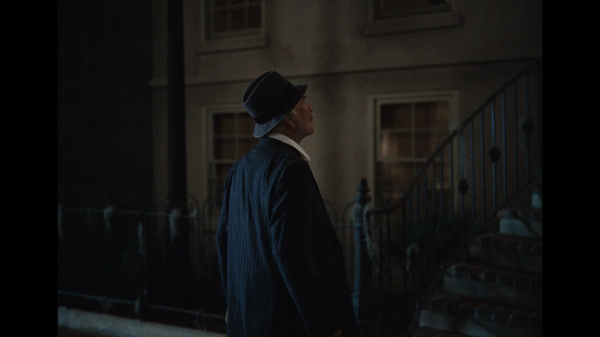 An elderly man in a suit and fedora hat standing outside at night near a staircase, looking up.