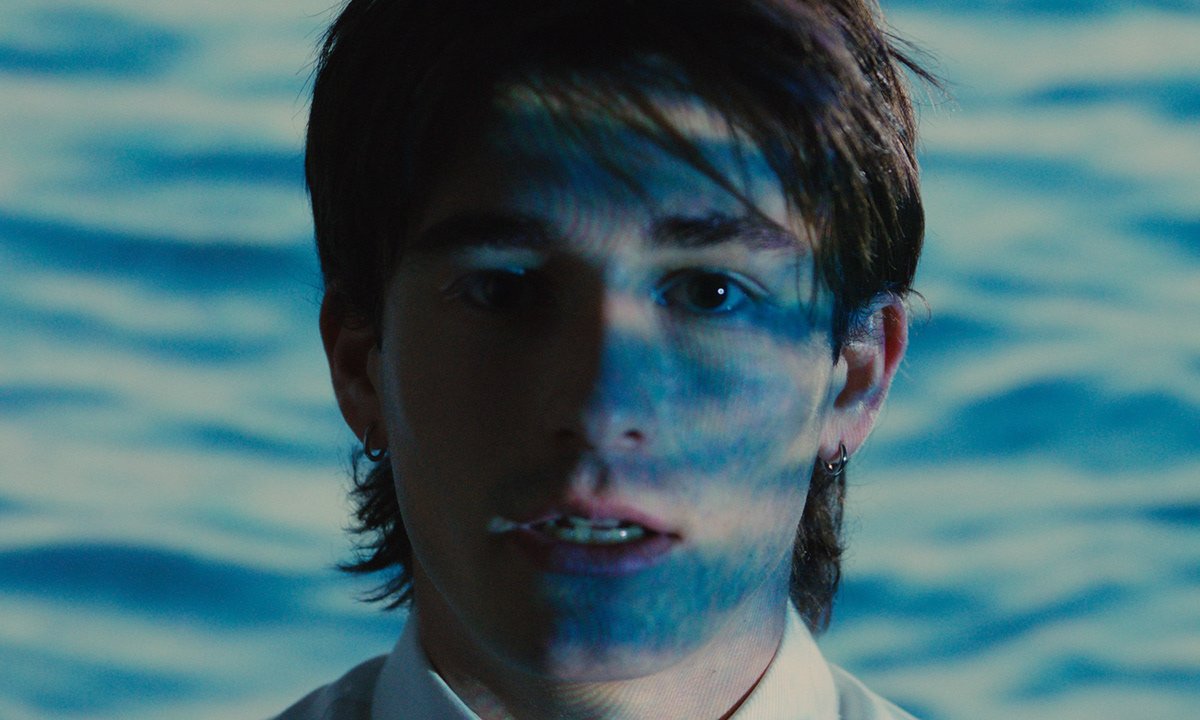 Close-up portrait of a young person with short dark hair, light skin, wearing earrings, eyes looking at the camera, partially shadowed face with an impression of water ripples reflected on it, set against a background of water.