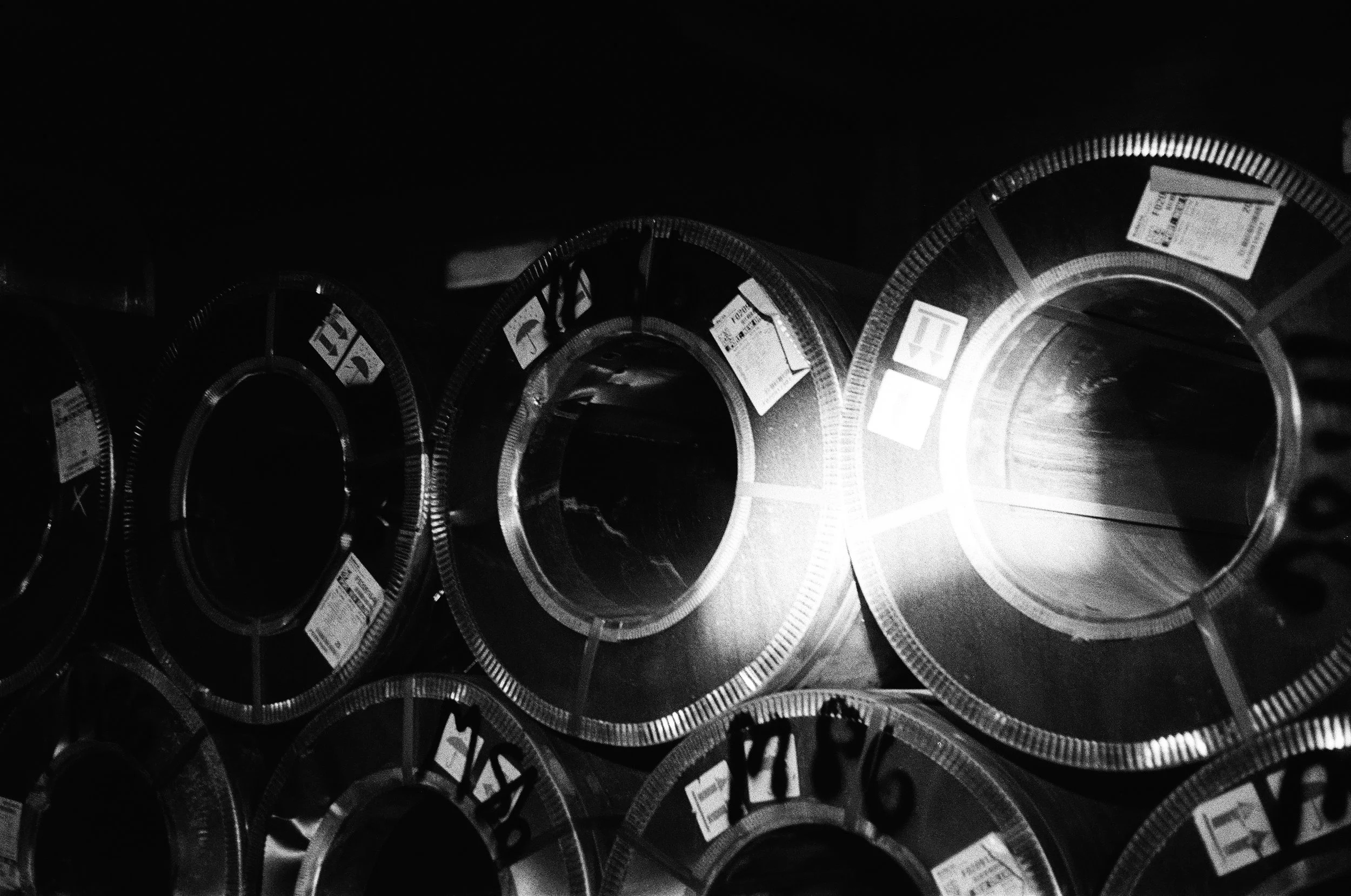 Stacks of metal pipes or cylinders stored on shelves in a dark industrial setting, with a light source causing reflections on their surfaces.