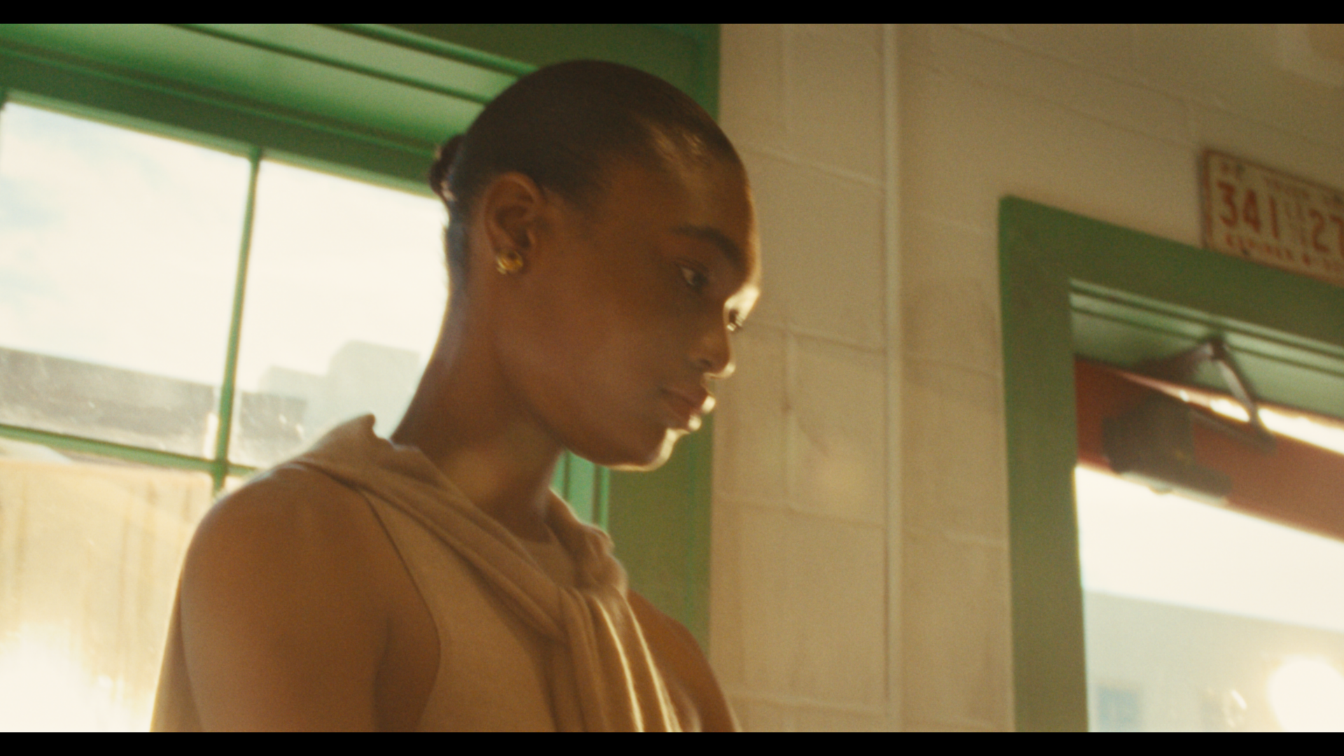 A woman with dark skin and dark hair tied back, wearing gold earrings and a sleeveless beige top, standing indoors near green window frames and a door, with sunlight streaming in.