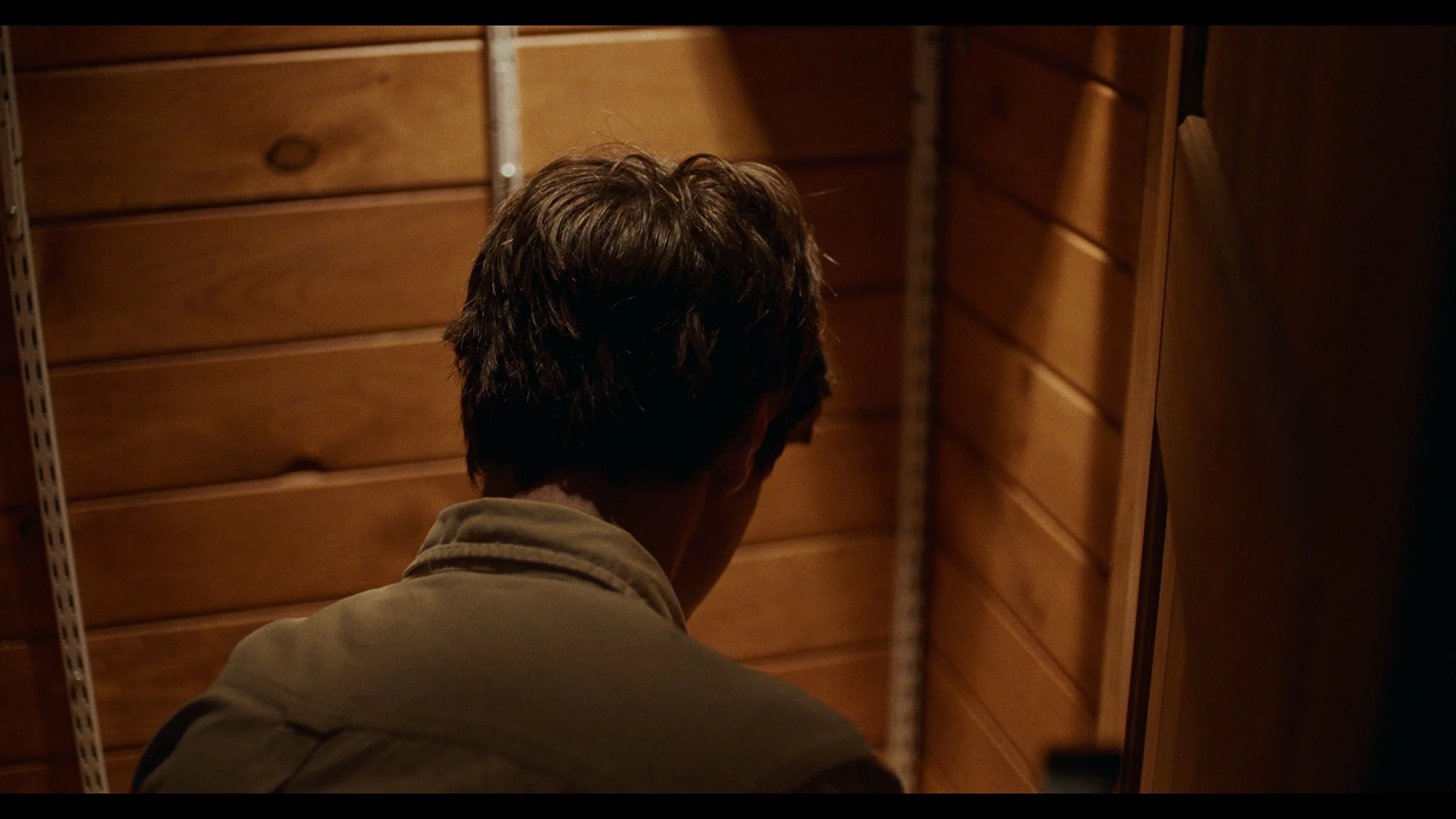 A person with dark hair is seen from behind, in a wooden-paneled room, facing away from the camera, wearing a jacket.