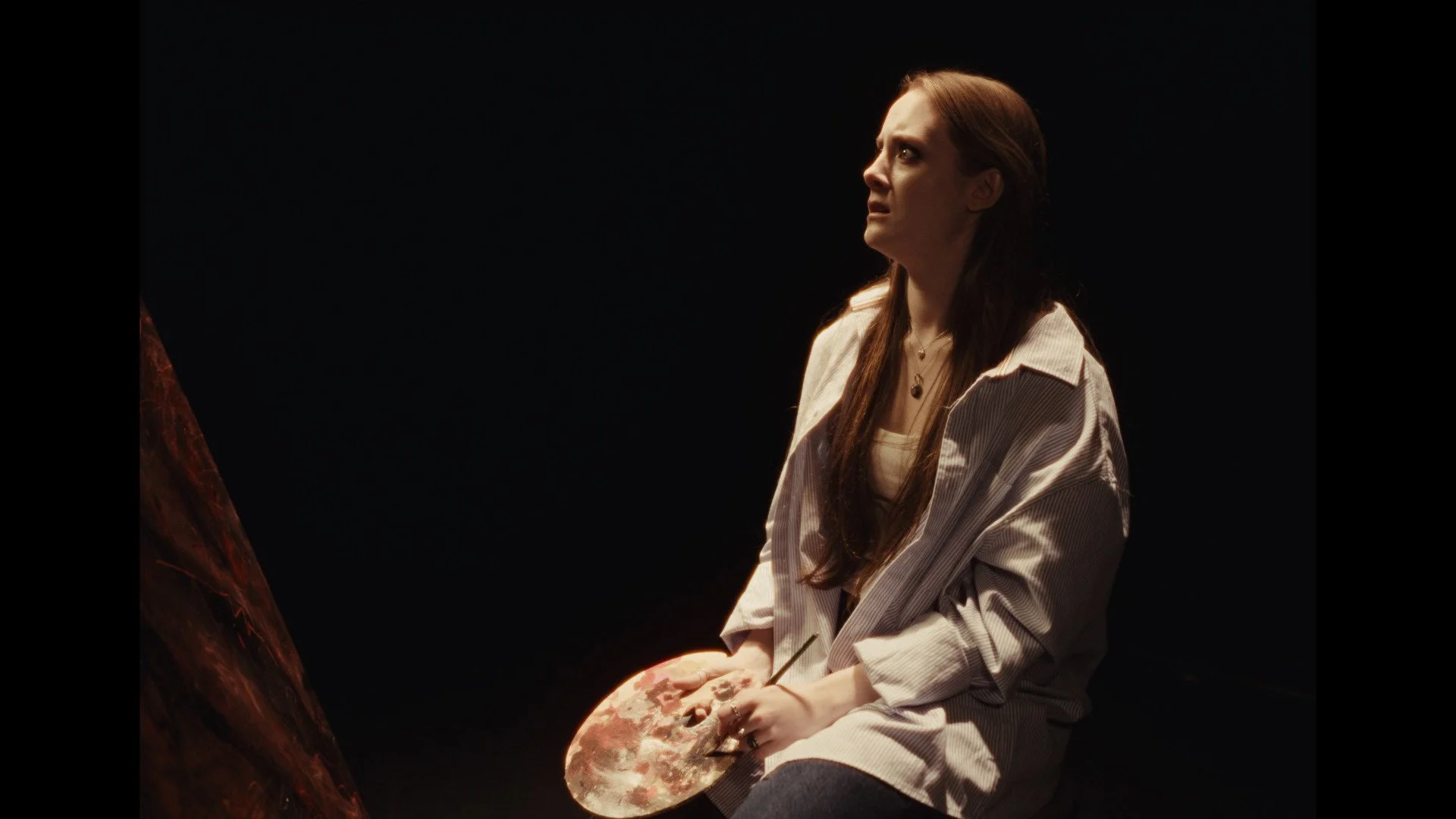A woman with long brown hair, wearing a beige jacket and white top, sitting in a dark room, holding a paint palette with earth tones, looking upward with a thoughtful expression.