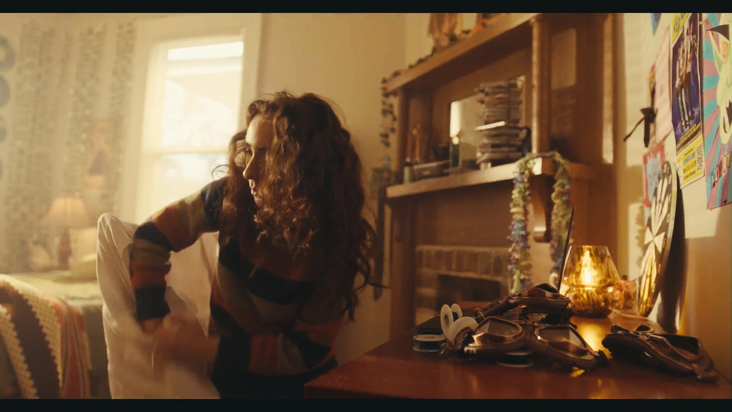 A woman with curly hair leaning over a table in a cozy, warmly lit room decorated with colorful artwork and a lamp, with shoes and accessories on the table. Mathias White: cinematographer and gaffer.