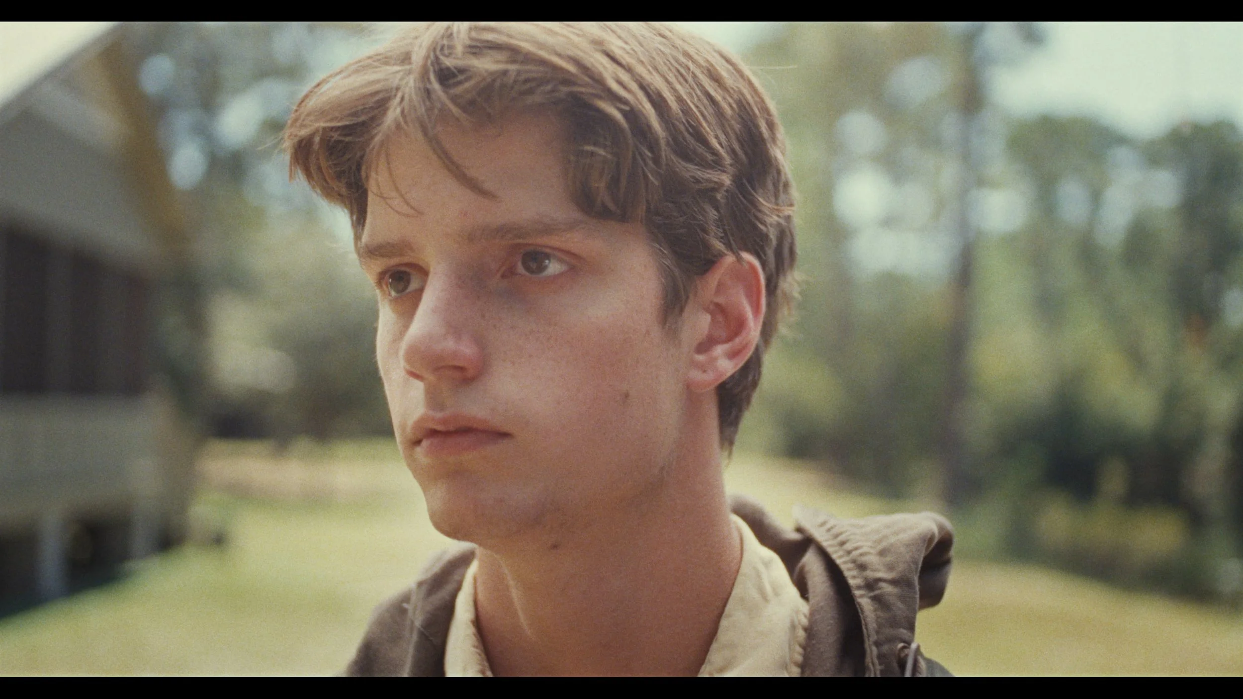 Close-up of a young man with brown hair and freckles looking pensively to the side outdoors, with a blurry background of trees and a house.