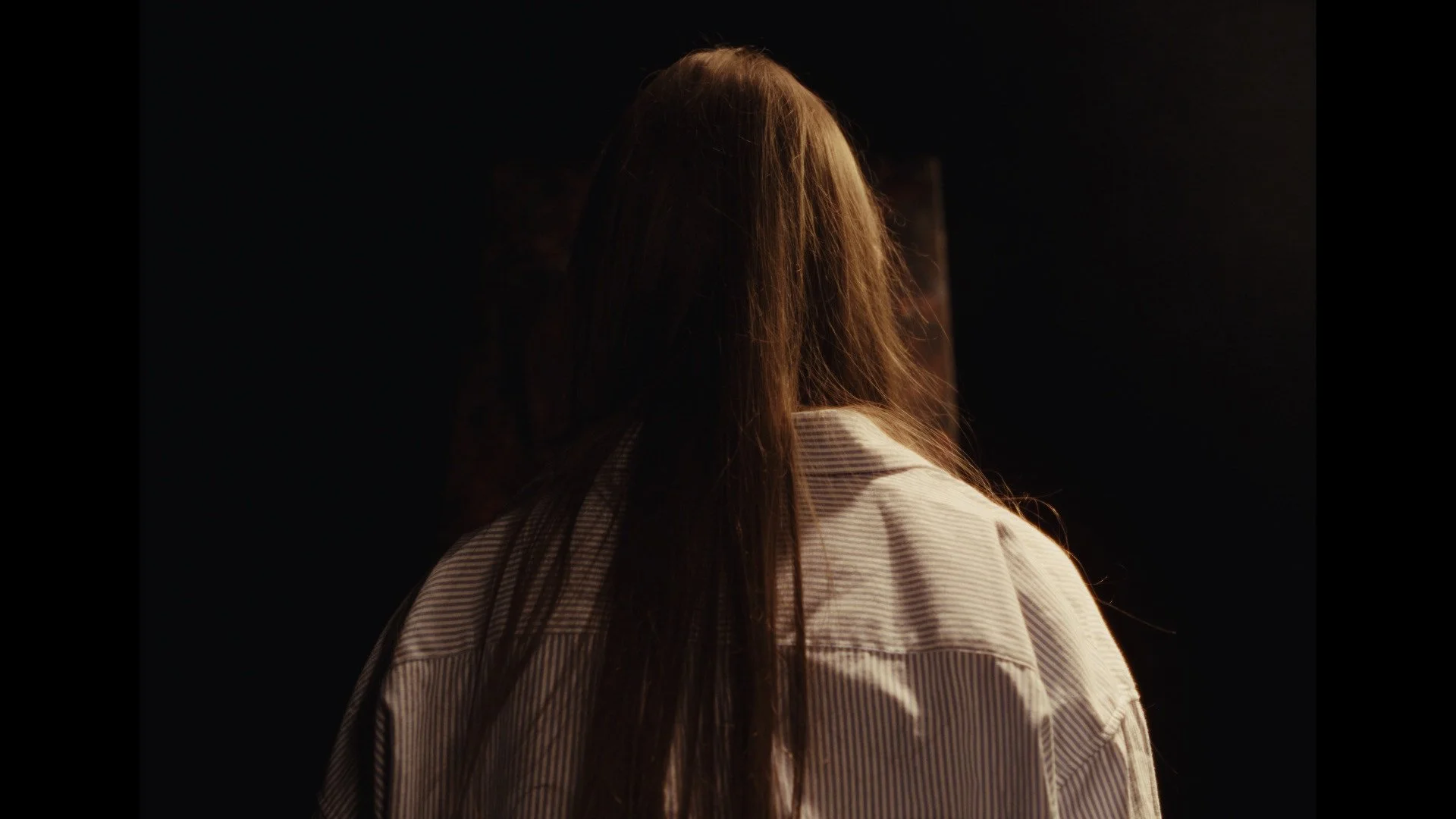 Back view of a woman with long hair wearing a light-colored, striped shirt, standing in a dark room with a faint glow on her hair.