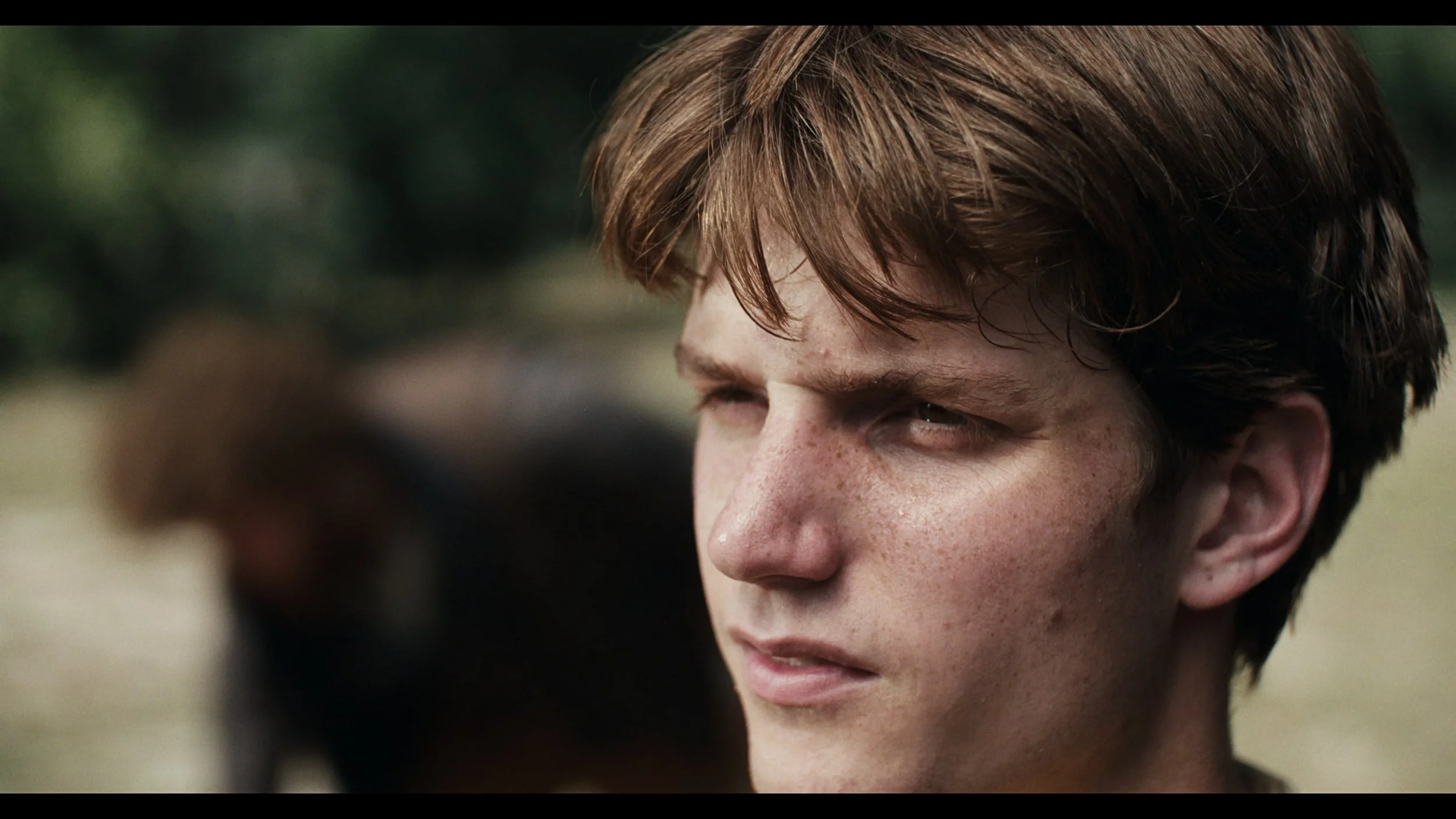 Close-up of a young man with short brown hair looking to the side outdoors, with a slightly blurred background.