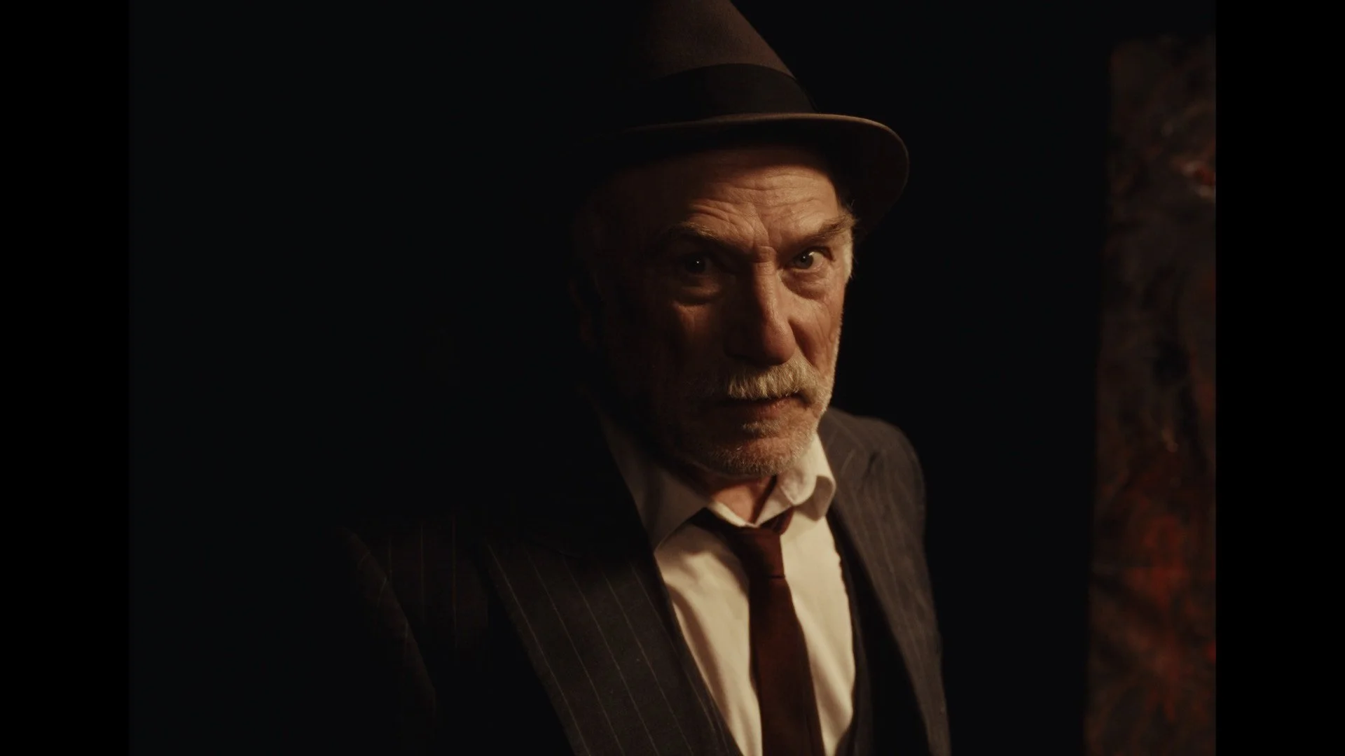 Close-up of an older man with a mustache wearing a fedora, pinstripe suit, white shirt, and tie, looking intensely at the camera in a dark setting.