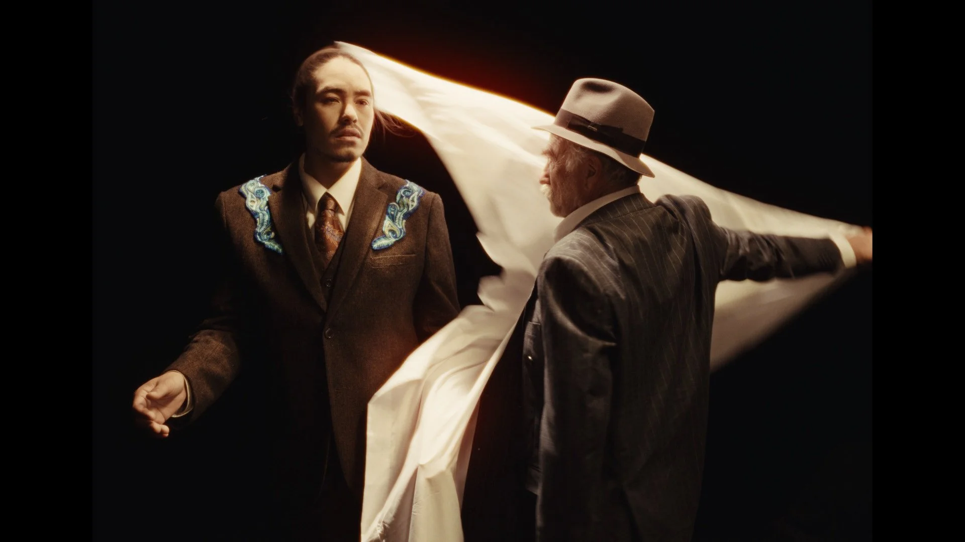 Two men in vintage suits, one with embroidered details, stand against a black background. One man with a hat is holding a large piece of fabric blocking the other man's face.