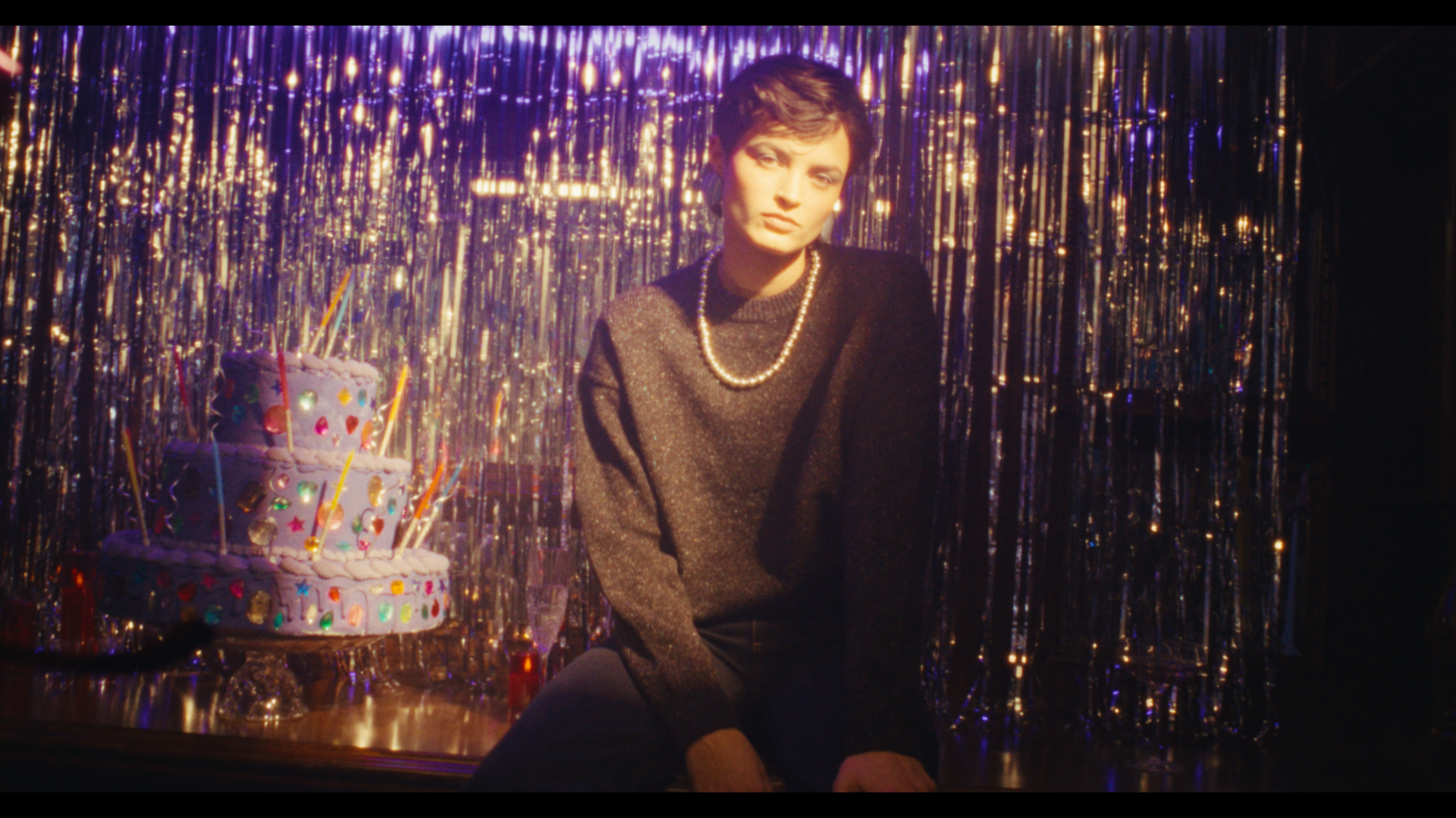 A woman with short dark hair, wearing a black sweater and pearl necklace, sitting in front of silver tinsel backdrop and a lit birthday cake with colorful decorations and candles.