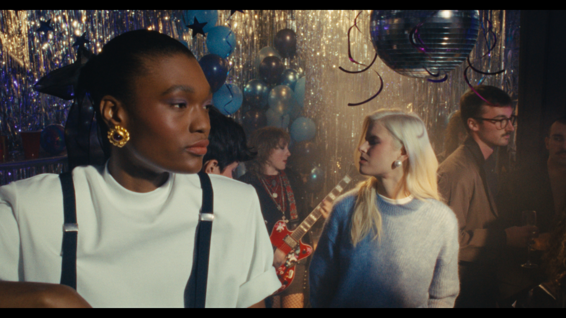 People at a party with balloons, streamers, and disco ball, some socializing and one person playing an electric guitar.