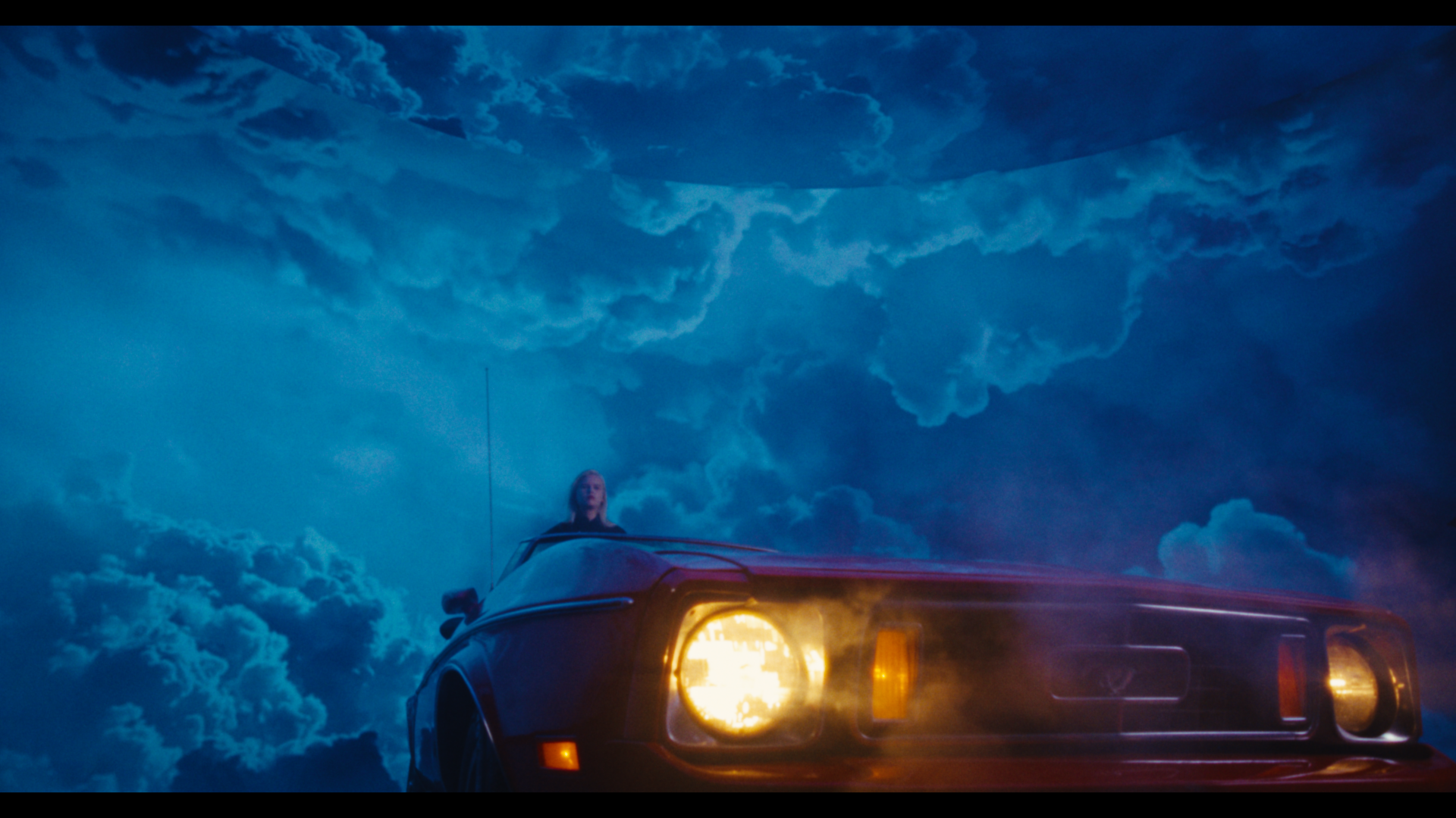 A woman sitting on the roof of a vintage car with the headlights on, under a dramatic cloudy sky with a cosmic or surreal atmosphere.