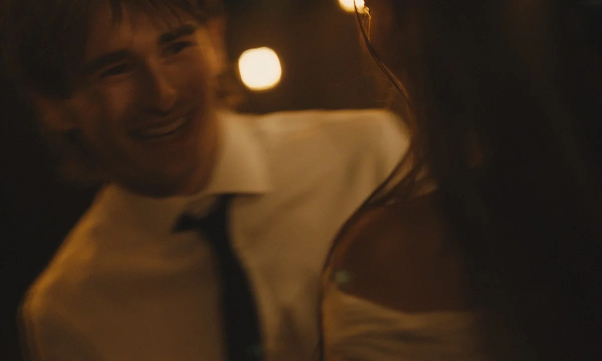 A person with curly hair and a smile, wearing a white shirt and dark tie, seen through a reflective surface, with warm lighting.