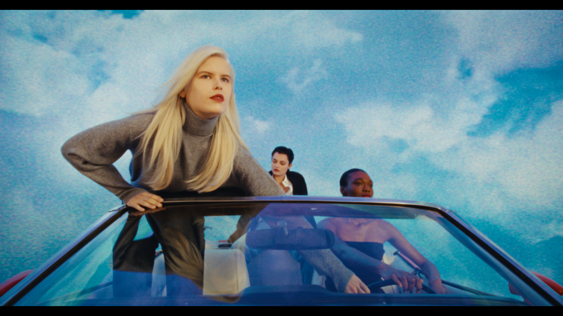 Three women on a convertible car under a cloudy sky, one woman leaning over the edge of the car looking forward, another woman driving, and a third woman sitting behind the driver.