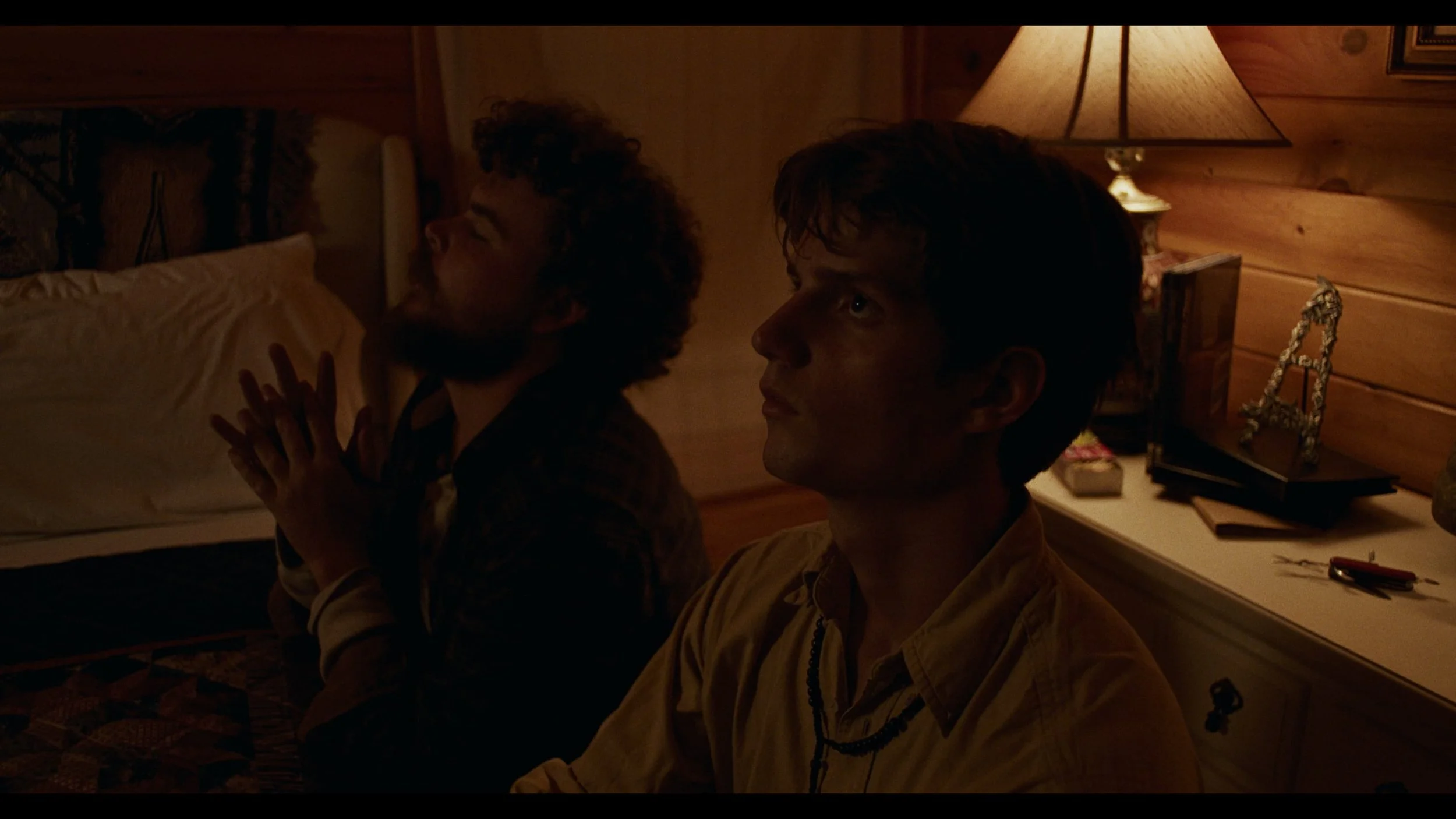 Two men sitting in a dimly lit room with wooden walls. One man is sitting on a bed with his eyes closed and hands together, appearing to pray or meditate. The other man is sitting nearby, looking serious, with a lamp and various objects on a dresser 