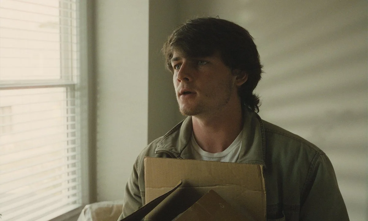 A young man with dark hair, wearing a beige jacket and white shirt, standing near a window with blinds, holding a cardboard box.