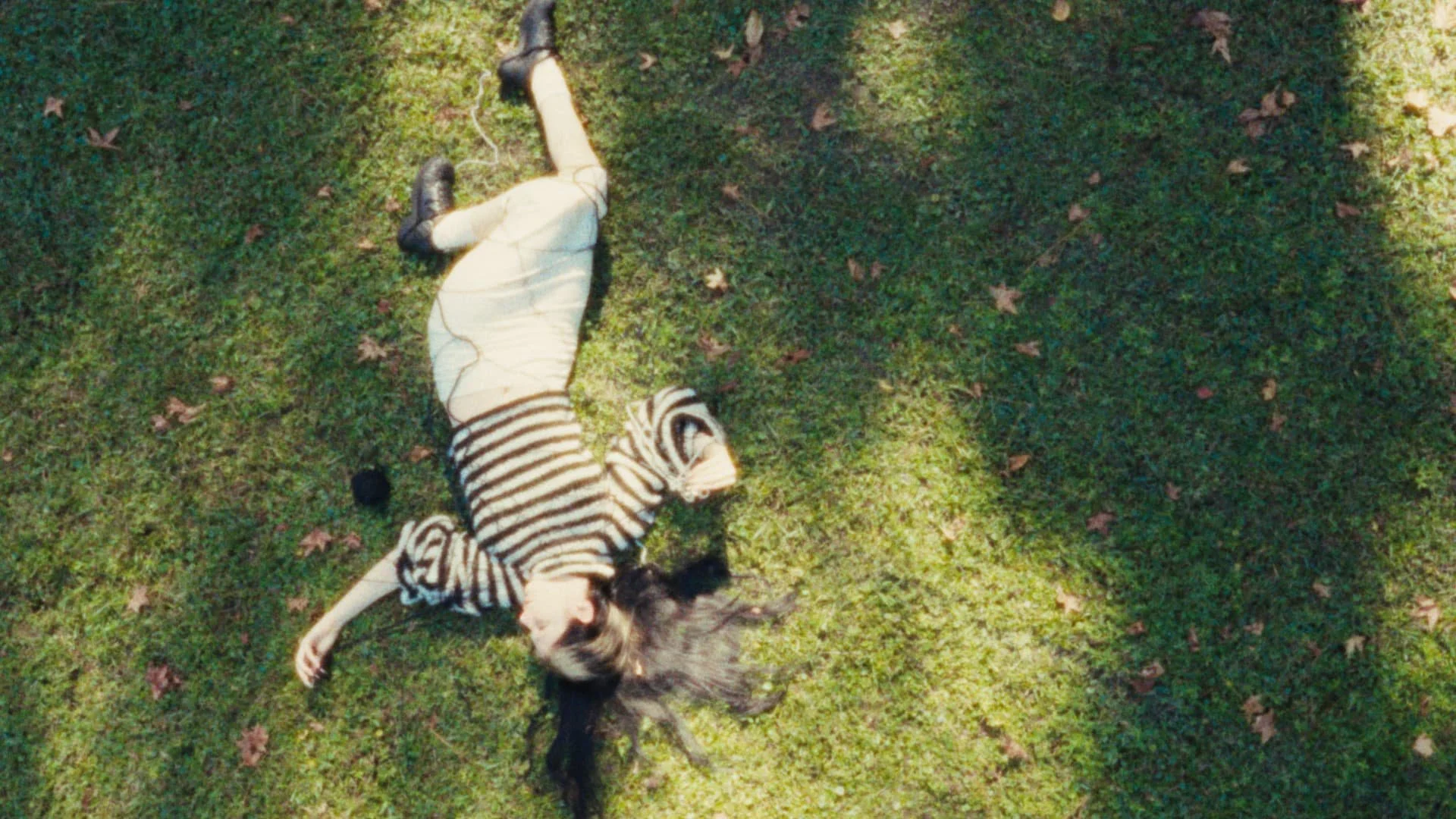 A girl in a striped sweater and beige pants is lying on grass, surrounded by fallen leaves, with her long hair spread out. She appears to be relaxing or resting.