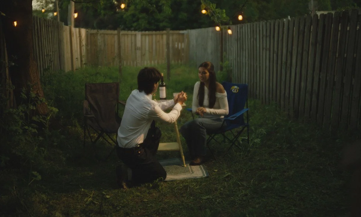 A man kneeling on a small rug in a backyard at night, proposing to a woman sitting in a camping chair, with string lights hanging overhead.