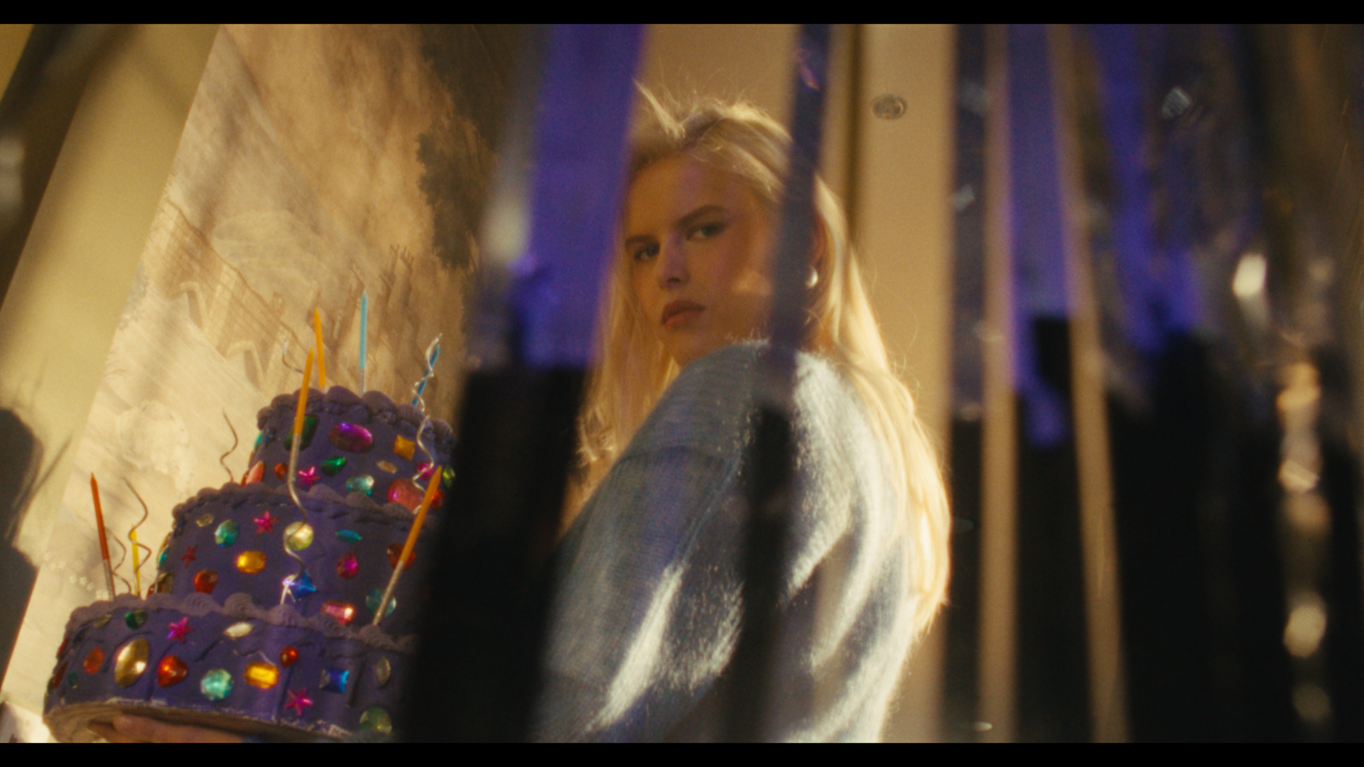 A blonde woman looking over her shoulder at the camera, standing next to a purple birthday cake decorated with colorful gems and candles, with a background of hanging metallic streamers.