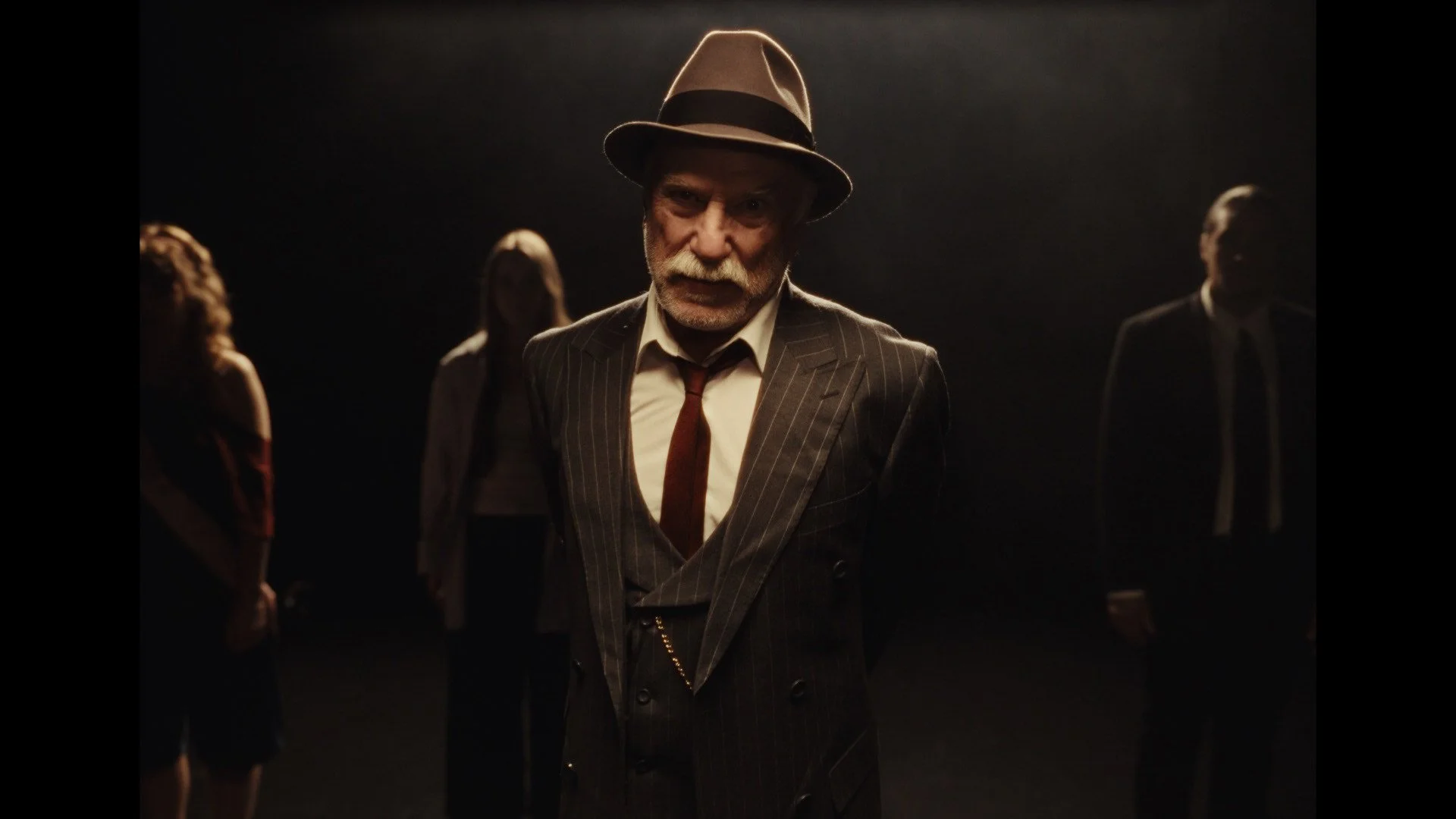 An elderly man with a white mustache and beard, wearing a pinstripe suit, a white shirt, a red tie, and a fedora, stands in a dark room with a spotlight on him, surrounded by four people in the background.