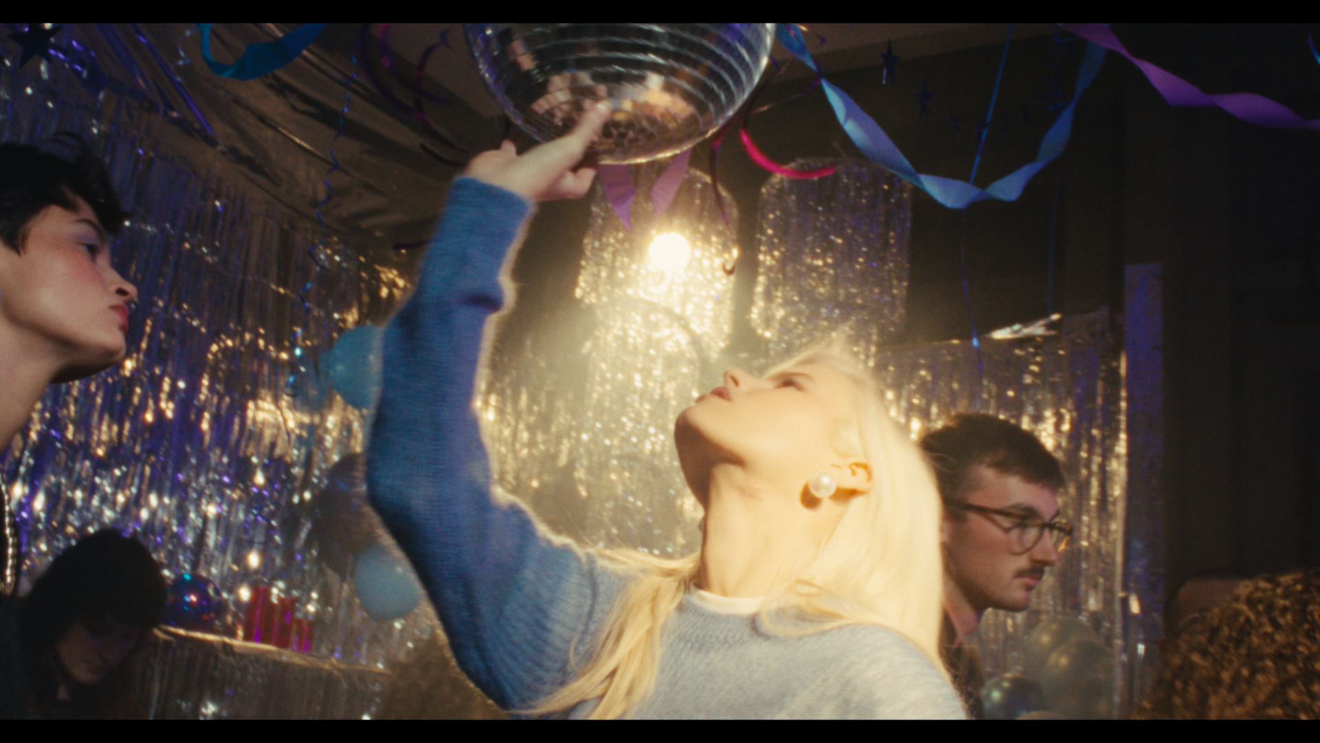 A young woman at a party reaching up to touch a disco ball surrounded by shiny decorations and streamers with other people in the background.