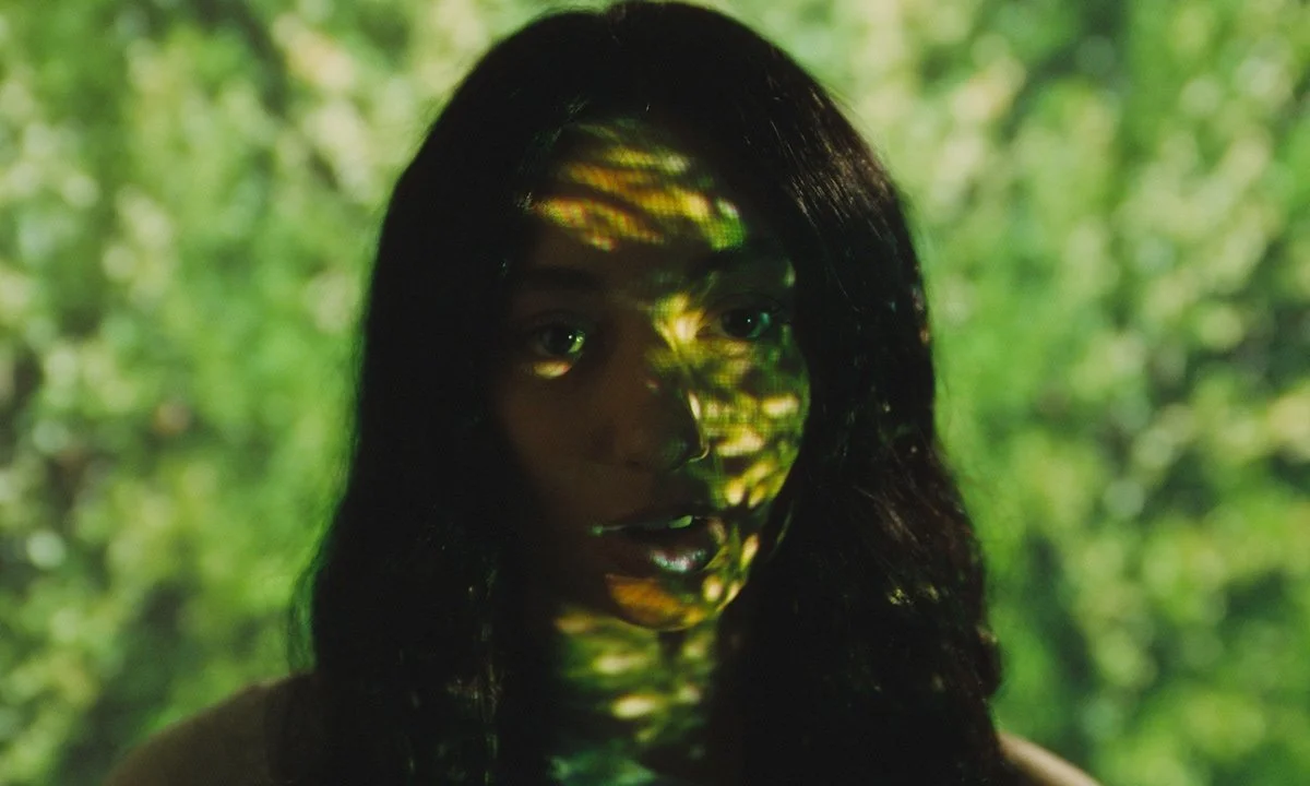 A woman with dark hair illuminated by green and yellow light patterns projected onto her face and hair, standing outdoors with a blurred green foliage background.