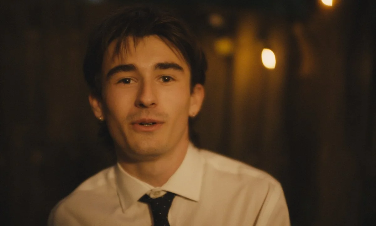 A young man with dark hair, wearing a white shirt and black tie, smiling in a dimly lit setting with blurred warm lights in the background.