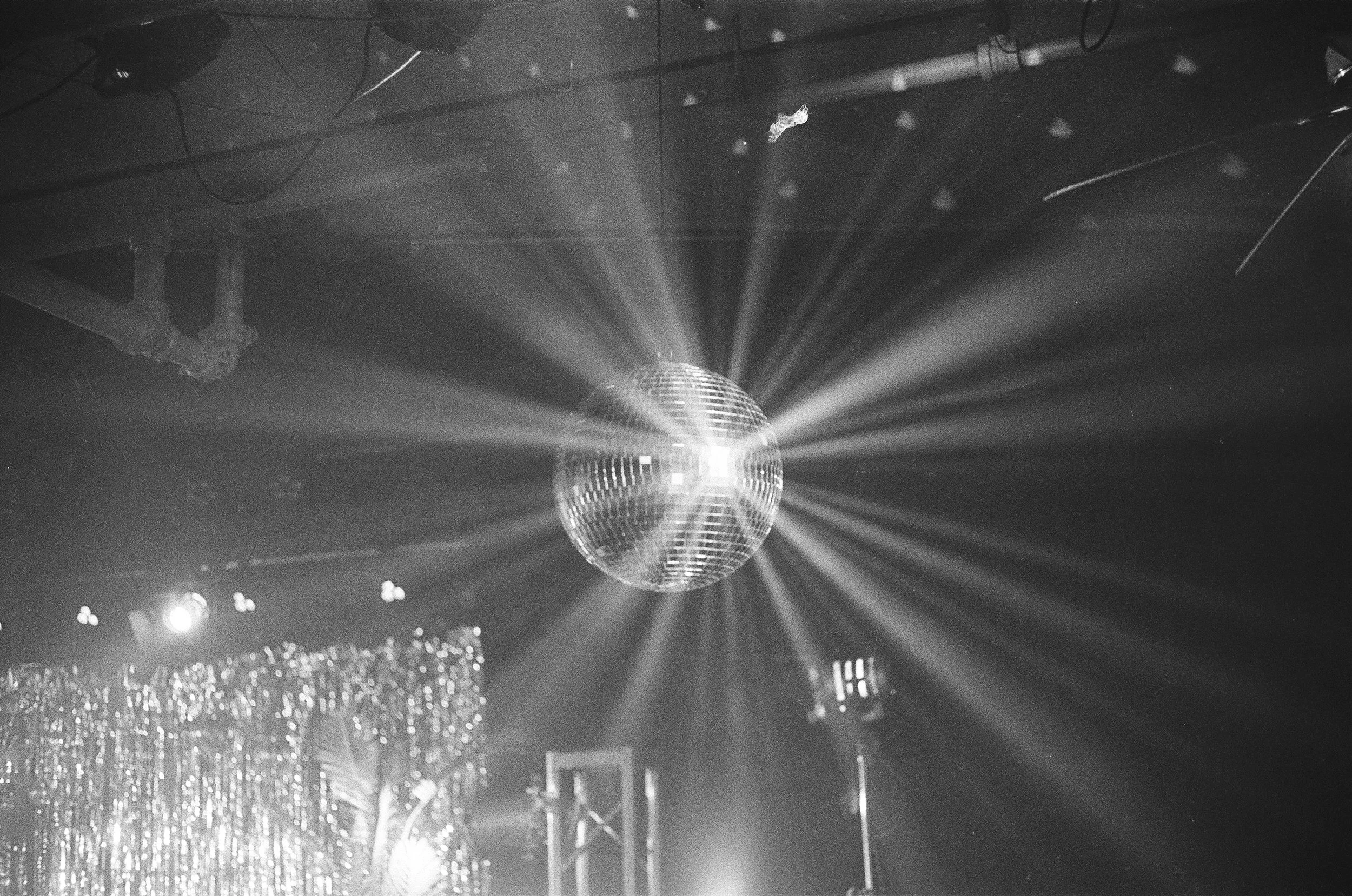A black and white photo of a disco ball hanging from the ceiling, reflecting light and creating beams of light radiating outward in a dark room with some stage lighting and decorations.
